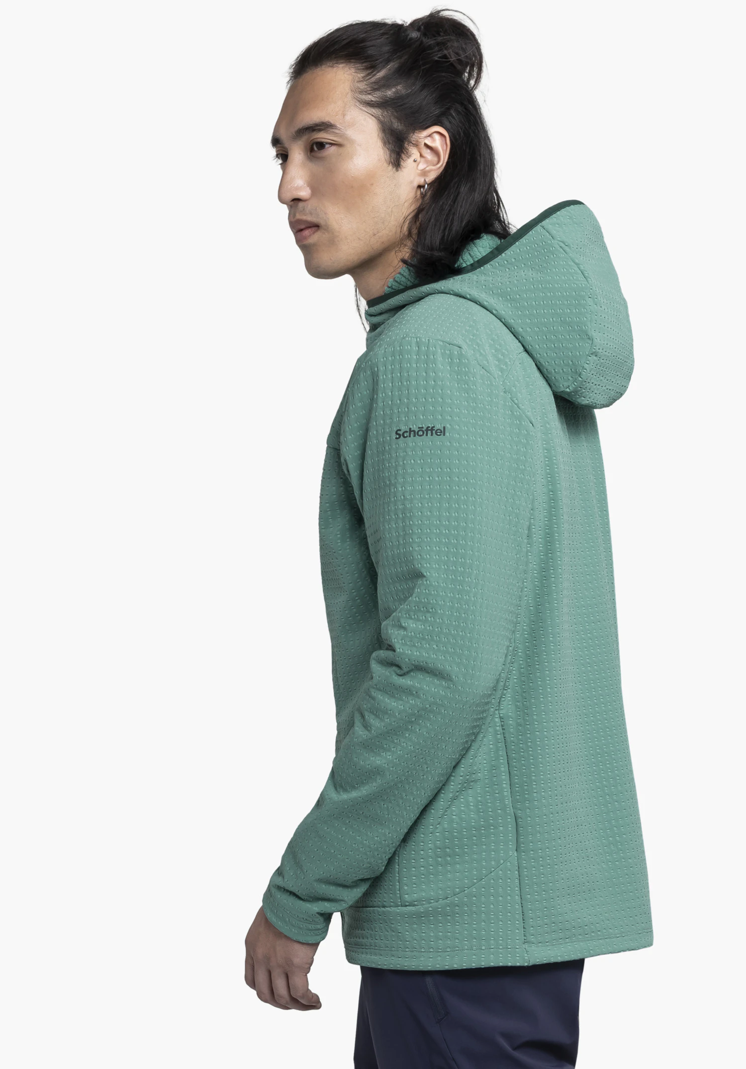 Fleece Hoody Style Enixa MEN : Un mannequin se tient de profil devant un fond neutre, mettant en valeur le sweat à capuche en vert. Le logo Schöffel discrètement placé sur le haut du bras souligne le look fonctionnel du vêtement.