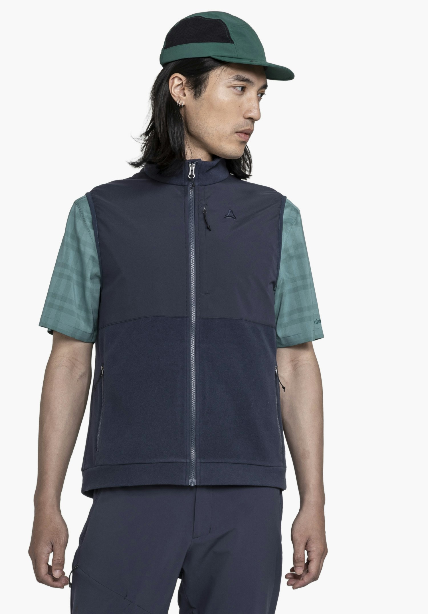 Fleece Vt Style Ash MEN on a model standing front-on and looking to the side. The blue gilet featuring a visible Schöffel-Logo on the left chest is worn over a green short-sleeved shirt, presented against a neutral background.