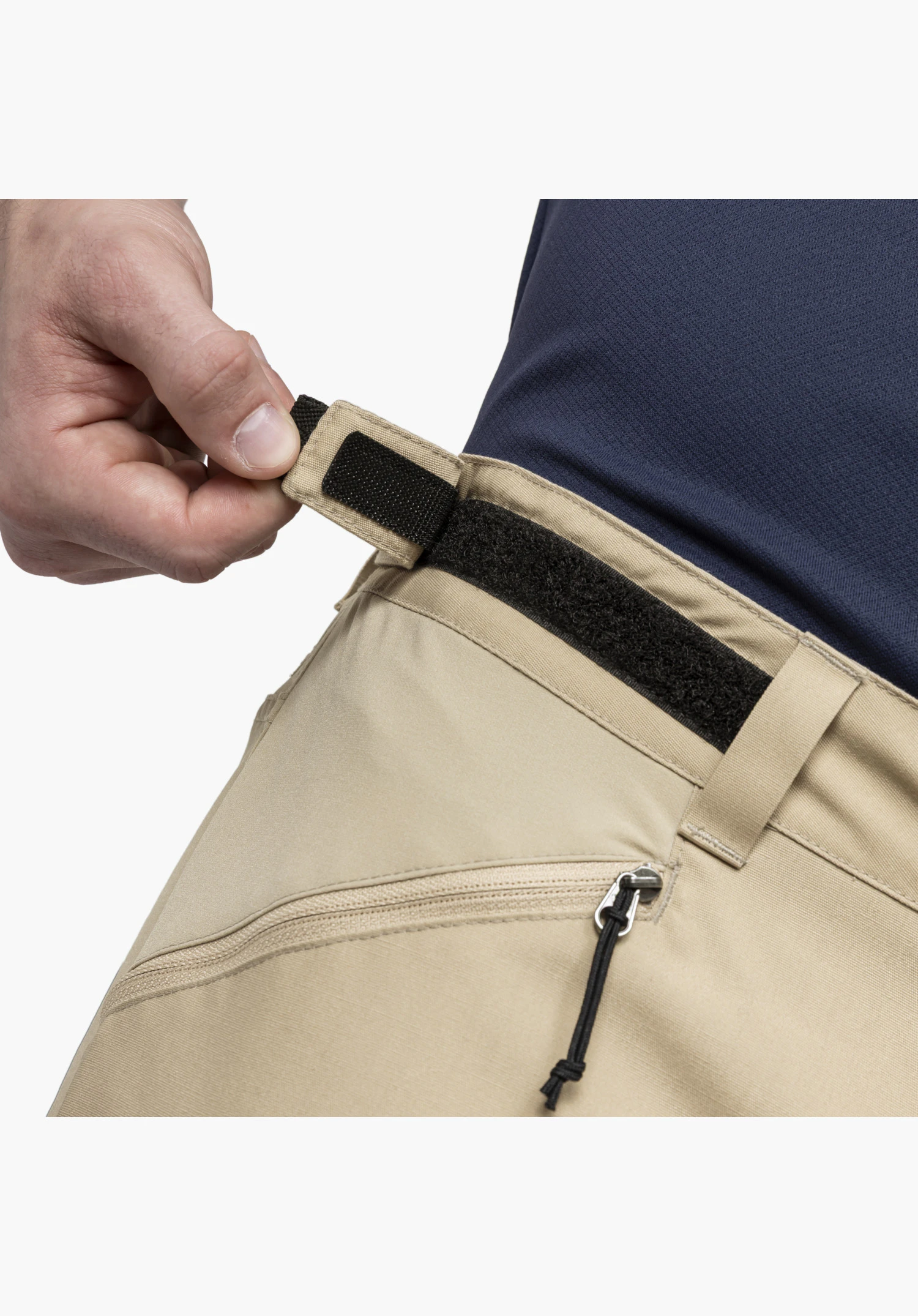Pants Style Maghera MEN in beige shows a detailed view of the adjustable waistband featuring a black hook-and-loop fastener being adjusted by a hand, positioned above a zipped pocket with a cord pull. Close-up against a white/neutral background.