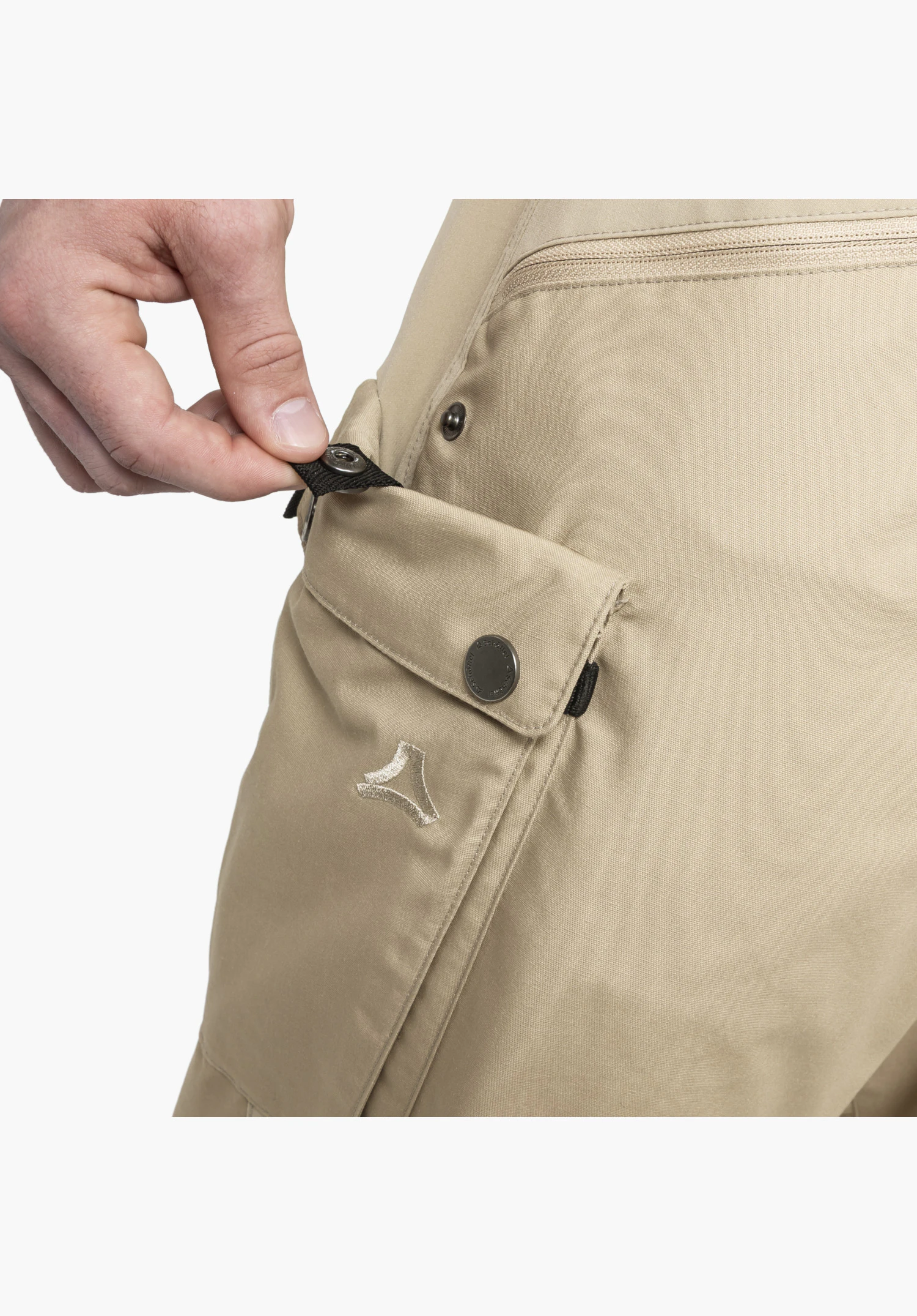 Pants Style Maghera MEN, detail view of the beige leg pocket where a hand is opening the popper fastening, featuring an embroidered Schöffel logo and a zip above, close-up against a white background.