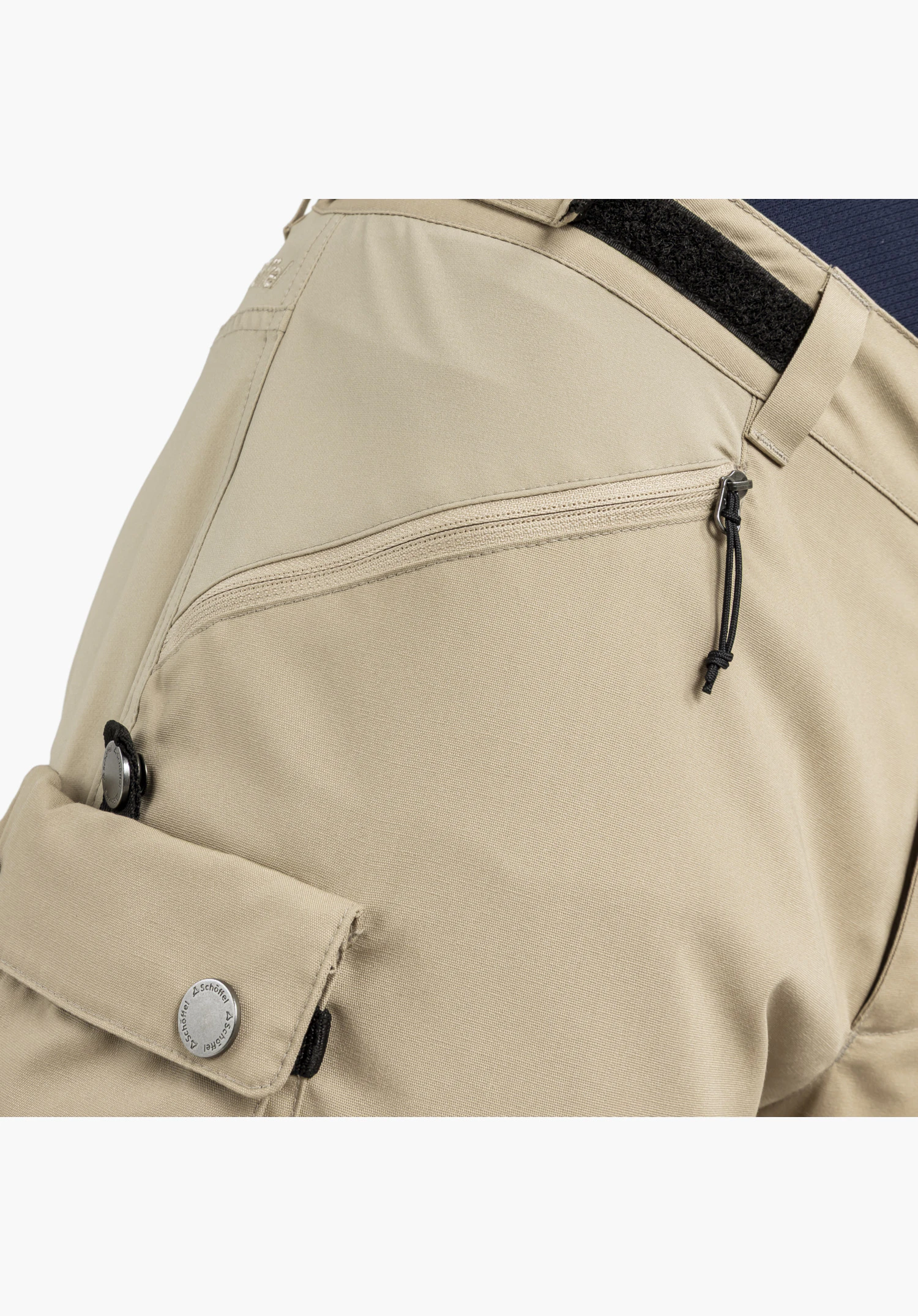Pants Style Maghera MEN en vue détaillée du tissu beige présentant une poche zippée en diagonale et une poche à rabat, fermée par un bouton-pression argenté gravé du Schöffel-Logo, gros plan sur fond blanc/neutre.