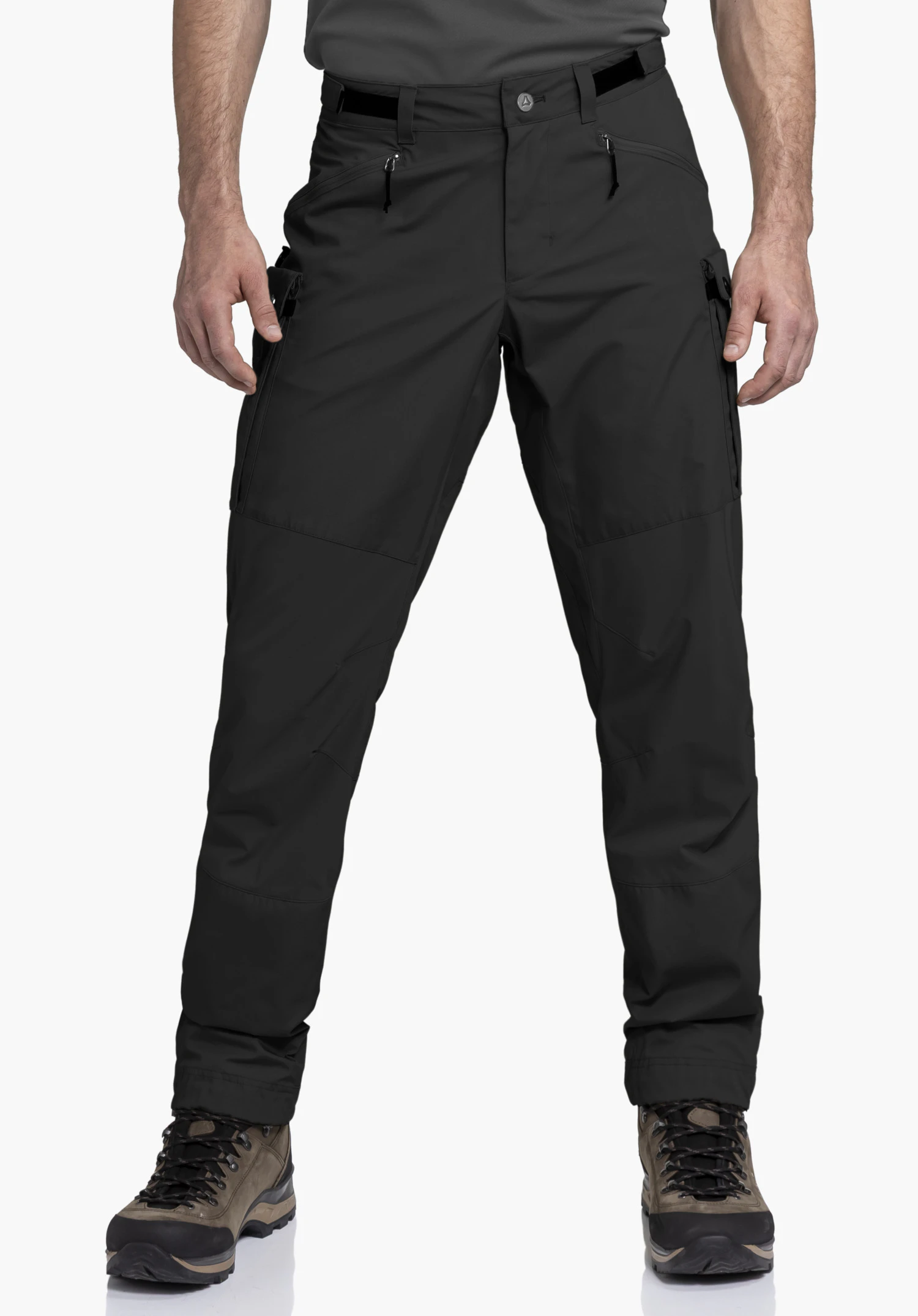 Pants Style Maghera MEN in black on a model. The model is standing frontally against a neutral background. This wear view shows the fit and the zip pockets of the trousers in combination with hiking boots.