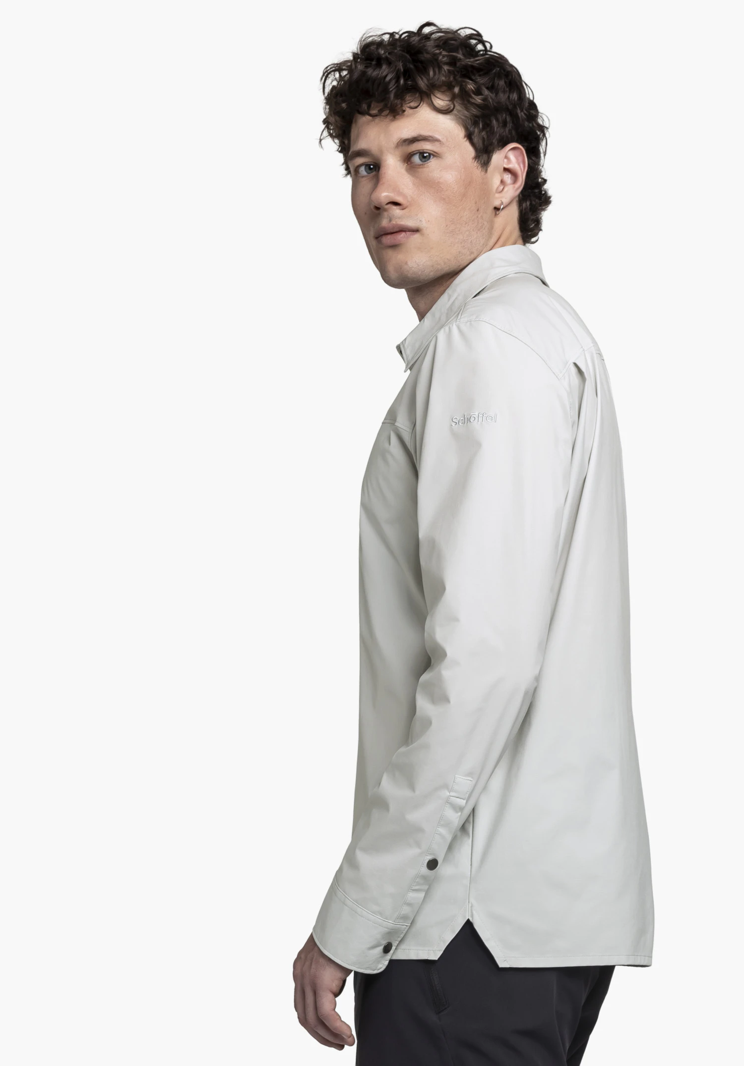 Shirt Style Maghera MEN in grey: A model is posing from a side angle against a neutral background, looking towards the camera. The long-sleeved shirt in grey features the subtle Schöffel logo on the upper arm, blending a relaxed fit with functional style.