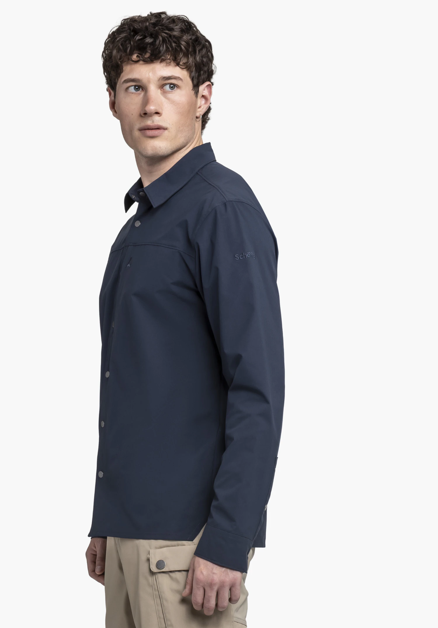 Shirt Style Maghera MEN en bleu. Un modèle présente la chemise à manches longues de profil sur un fond neutre. Le regard du modèle est tourné vers le côté, mettant en valeur le logo Schöffel discret sur la manche.