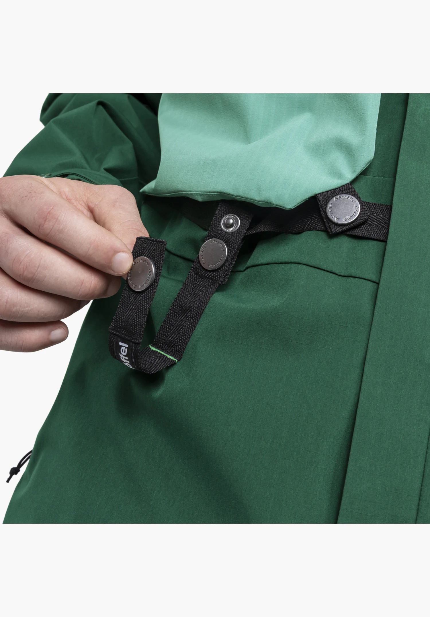 2L Jacket Style Maghera MEN zeigt im Detail eine Hand, die ein schwarzes Befestigungsband mit metallenen Druckknöpfen und Schöffel-Logo auf dem grünen Stoff greift, Nahaufnahme vor weißem/neutralem Hintergrund.