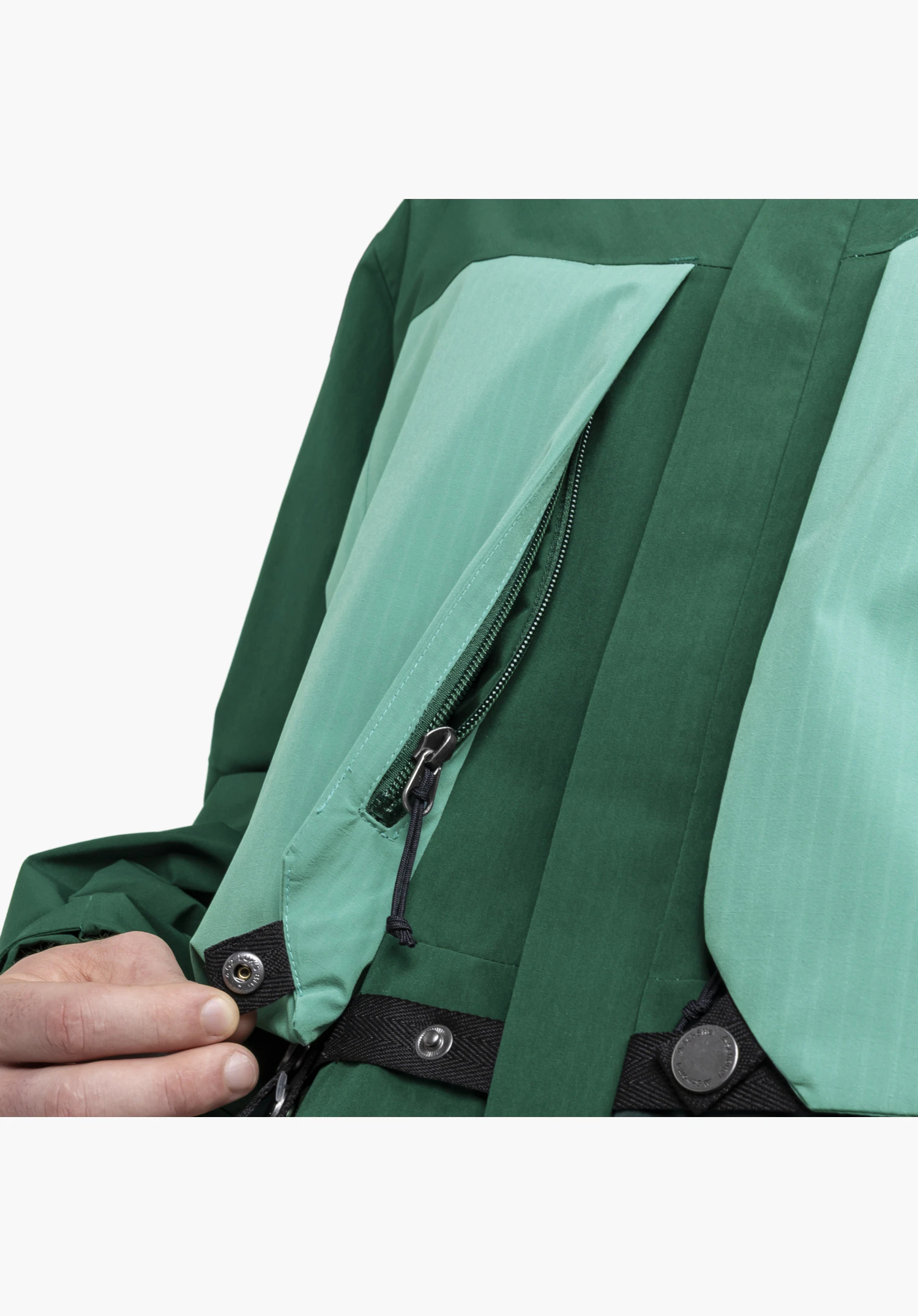 2L Jacket Style Maghera MEN: Detailansicht der zweifarbigen grünen Stoffpartien mit geöffnetem Reißverschluss, Kordelzug und schwarzen Riemen mit eingraviertem Schöffel-Logo auf den Metalldruckknöpfen, wovon einer von einer Hand gehalten wird. Nahaufnahme vor weißem Hintergrund.