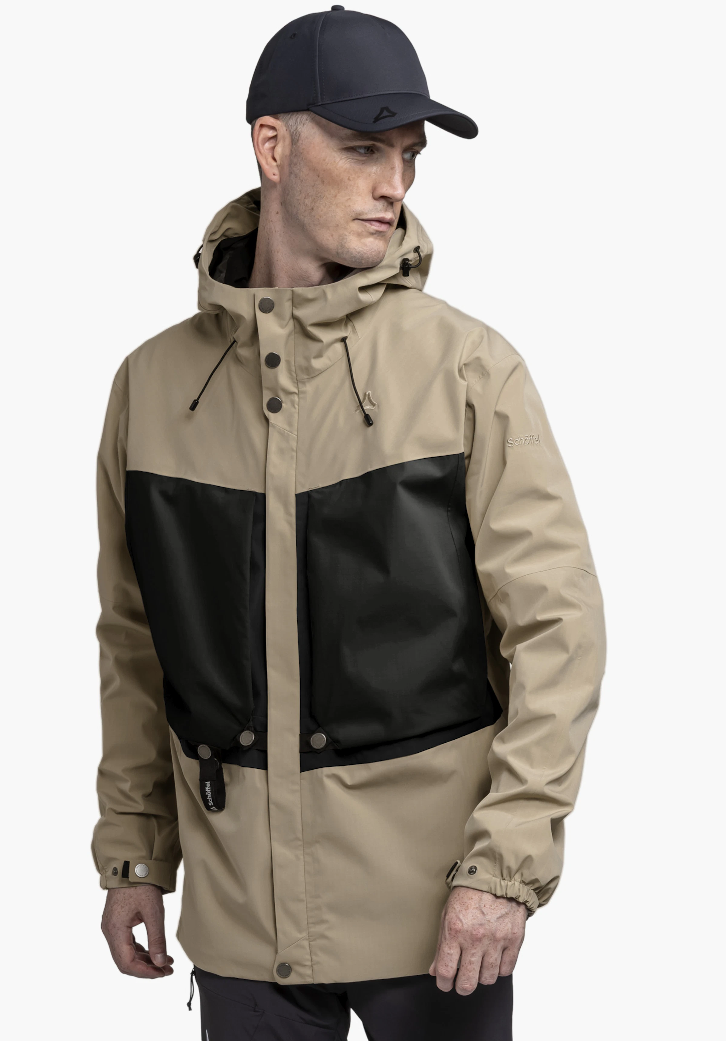 2L Jacket Style Maghera MEN. A model stands slightly to the side wearing the beige jacket with striking black inserts. The Schöffel logo is visible on the chest and the left sleeve, photographed against a neutral background.