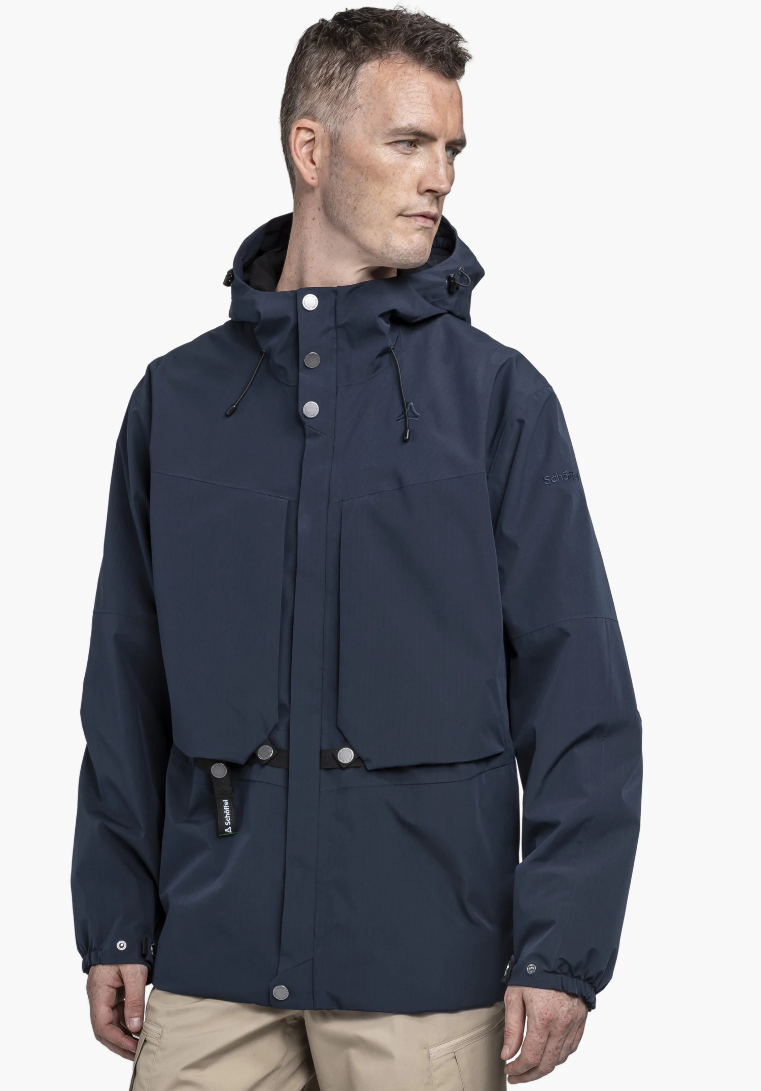 2L Jacket Style Maghera MEN. Ein Model trägt die blaue Jacke, steht leicht seitlich, blickt zur Seite und präsentiert markante Details wie die großen Vordertaschen, silbernen Knöpfe sowie das dezente Schöffel-Logo vor einem neutralen Hintergrund.
