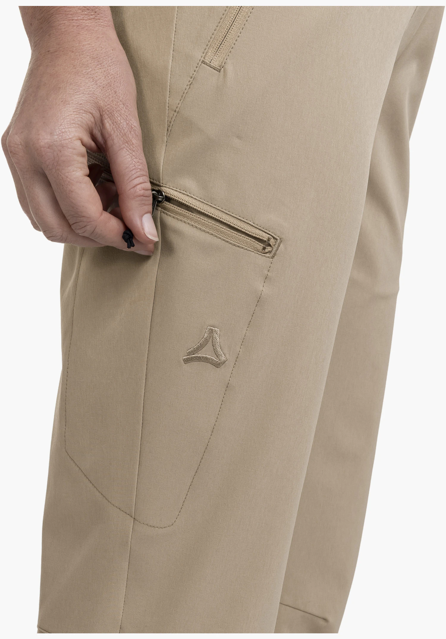 Pants Style Chavuma WMN mit Fokus auf die seitliche Reißverschlusstasche, die von einer Hand geöffnet wird, sowie das gestickte Schöffel-Logo auf dem beigen Stoff, Nahaufnahme vor weißem Hintergrund.