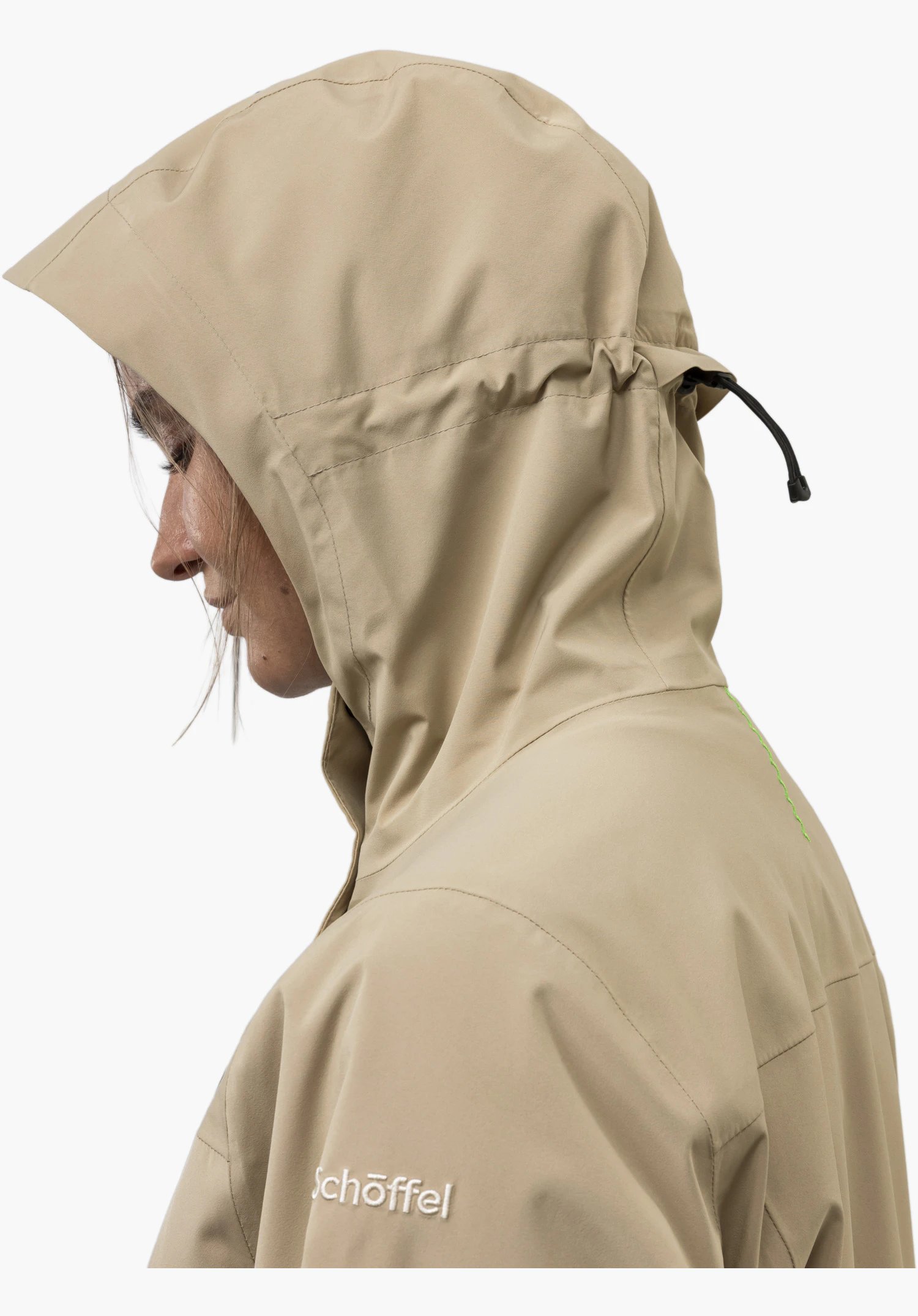 CIRC Jacket Style Smue WMN mit Fokus auf das gestickte weiße Schöffel-Logo auf dem beigen Stoff, den schwarzen Kordelzug an der Kapuze und das auffällige grüne Zickzack-Designelement, Nahaufnahme vor weißem/neutralem Hintergrund.