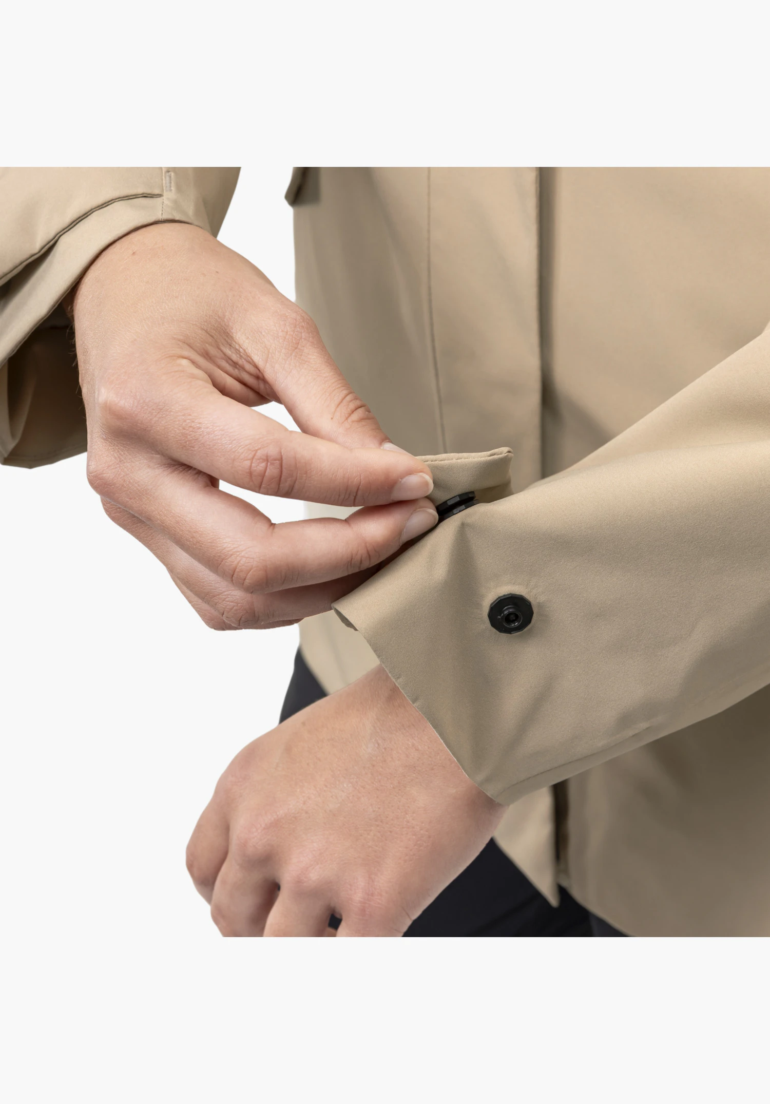 CIRC Jacket Style Smue WMN, Detail des beigen Ärmelbündchens mit einer Hand, die den schwarzen Druckknopf schließt, als Nahaufnahme vor weißem/neutralem Hintergrund.