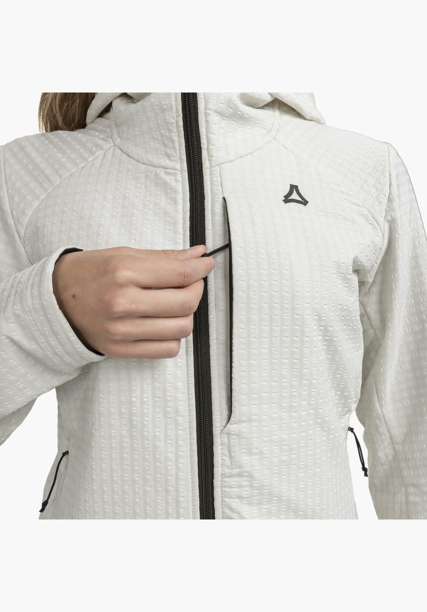 Fleece Hoody Style Enixa WMN präsentiert die waffelartige Struktur des naturfarbenen Stoffs im Brustbereich. Eine Hand bedient den schwarzen Reißverschluss der Brusttasche, neben dem sich das farblich abgesetzte Schöffel-Logo befindet. Nahaufnahme vor weißem Hintergrund.