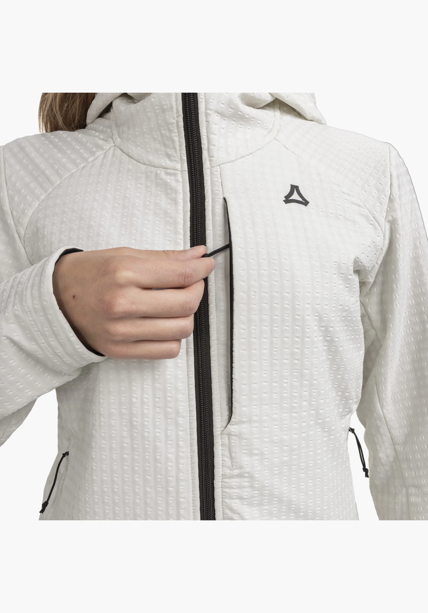 Fleece Hoody Style Enixa WMN präsentiert die waffelartige Struktur des naturfarbenen Stoffs im Brustbereich. Eine Hand bedient den schwarzen Reißverschluss der Brusttasche, neben dem sich das farblich abgesetzte Schöffel-Logo befindet. Nahaufnahme vor weißem Hintergrund.