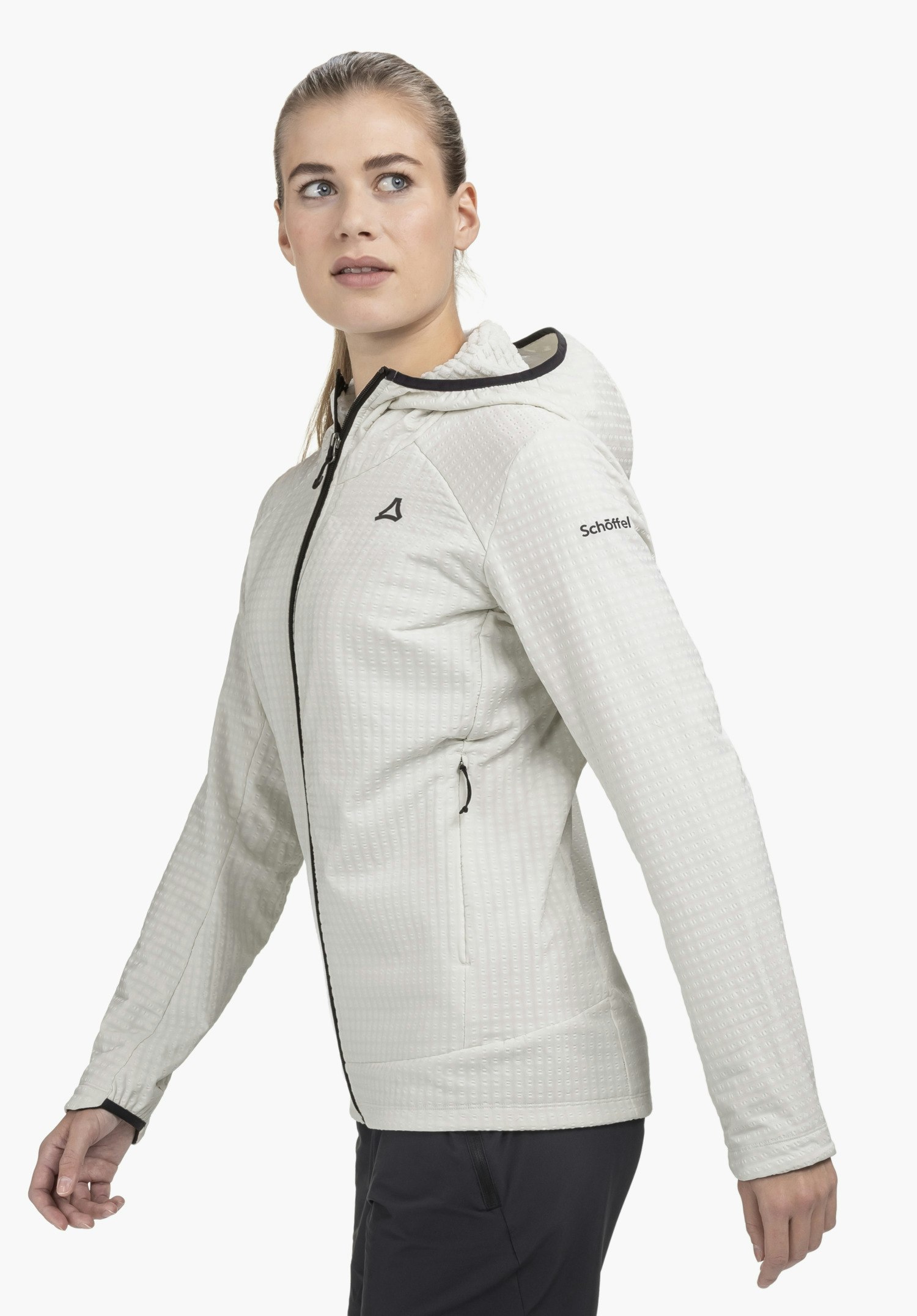 Fleece Hoody Style Enixa WMN in der Farbe Natur, getragen von einem Model, das leicht seitlich in einer dynamischen Pose steht. Zu sehen sind der schwarze Frontreißverschluss, schwarze Details an der Kapuze sowie das Schöffel-Logo auf der Brust und am Ärmel vor einem neutralen Hintergrund.