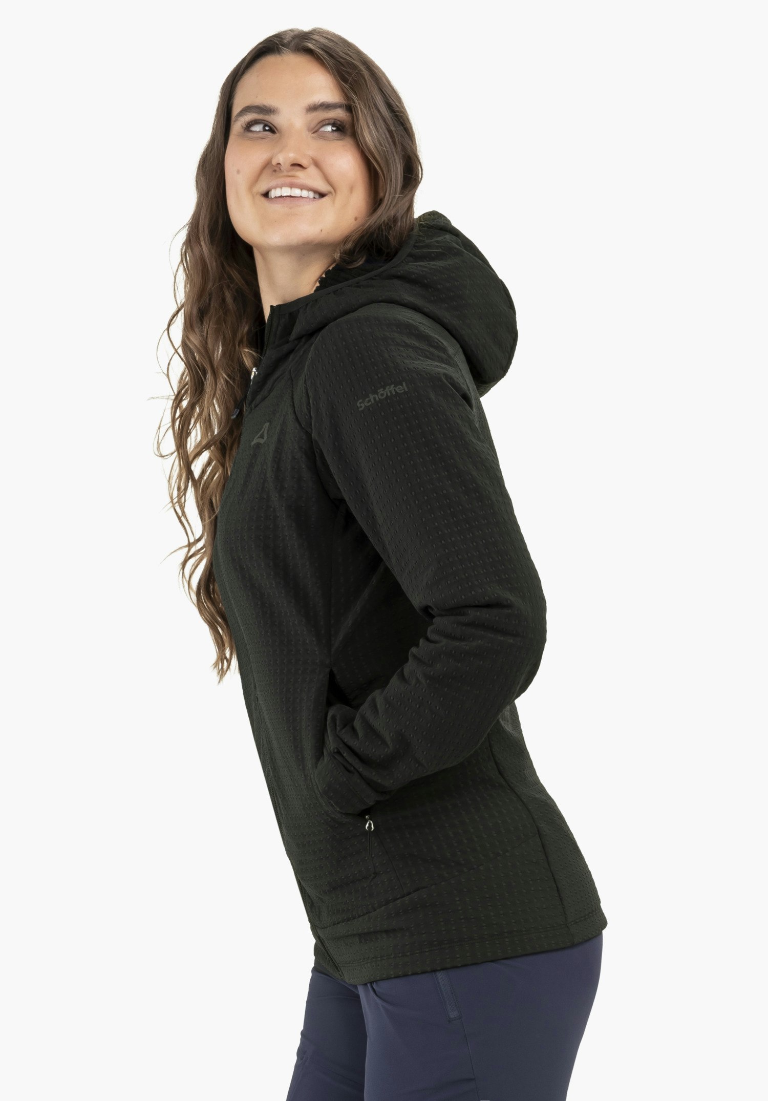 Fleece Hoody Style Enixa WMN, getragen von einem Model, das seitlich steht und lächelnd über die Schulter blickt. Sie hat eine Hand in der Tasche des schwarzen Hoodys, auf dessen Oberarm das Schöffel-Logo sichtbar ist, vor einem neutralen Hintergrund.