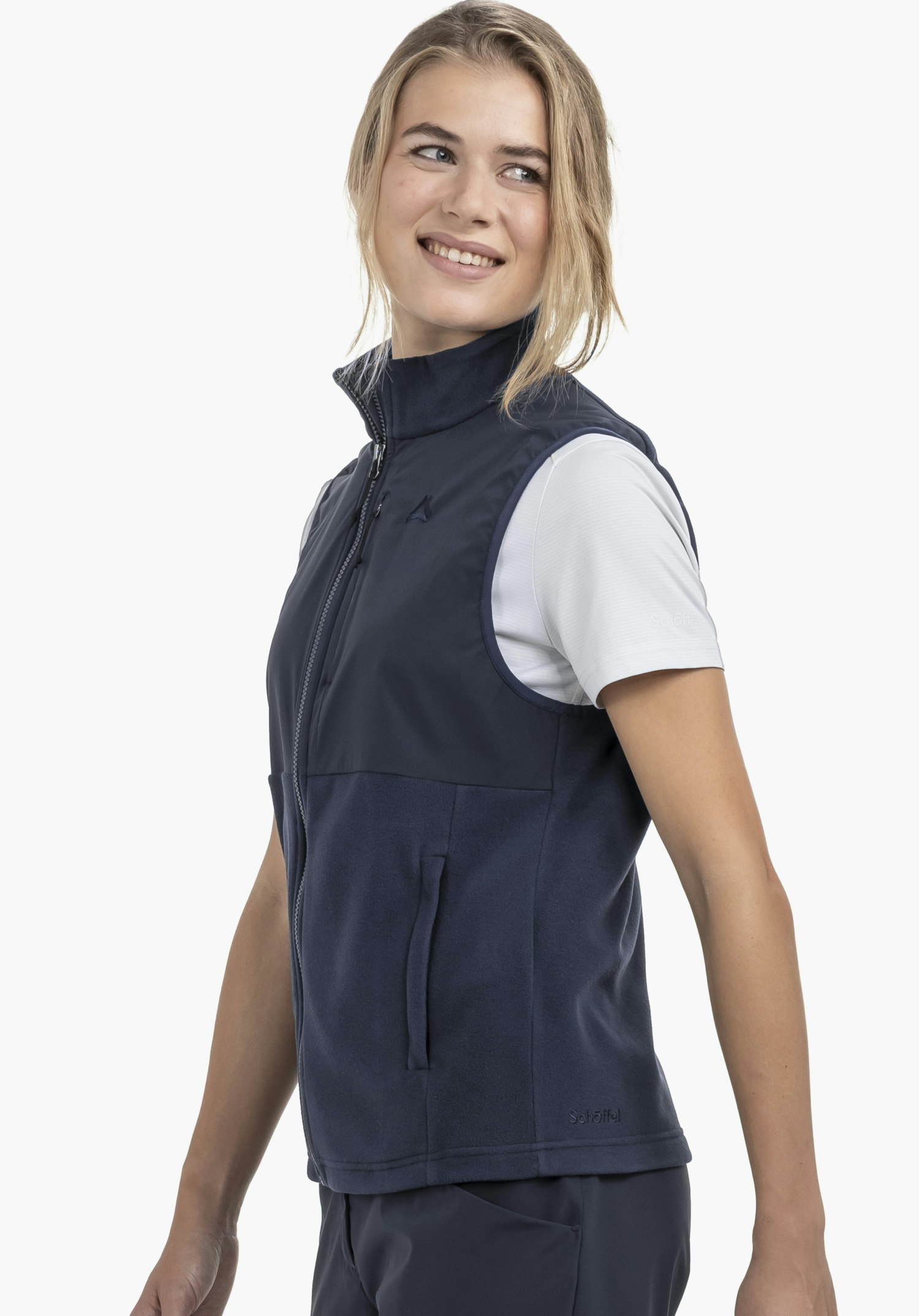 Fleece Vt Style Ash WMN worn by a model standing sideways and smiling. The blue gilet features a full-length front zip, a side pocket, and the Schöffel logo on the left chest and lower hem. Photographed against a neutral background.