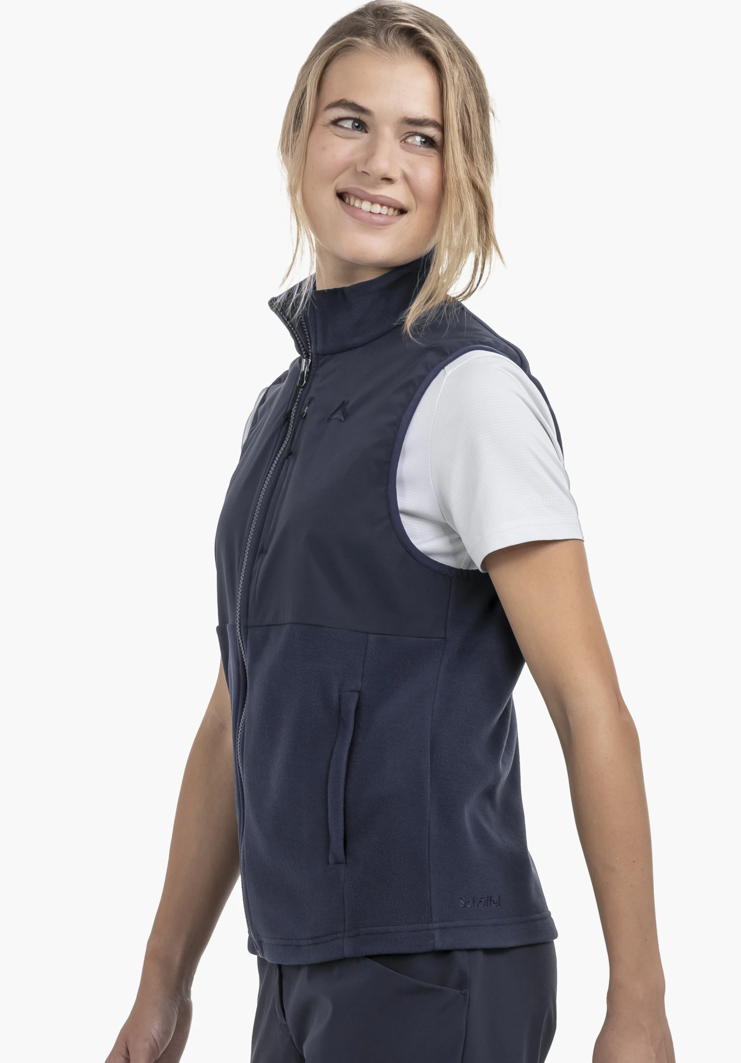 Fleece Vt Style Ash WMN gedragen door een model dat zijwaarts staat en glimlacht. De blauwe bodywarmer toont een doorlopende ritssluiting aan de voorkant, een zijzak en het Schöffel-logo op de linkerborst en de onderste zoom. Gepresenteerd tegen een neutrale achtergrond.