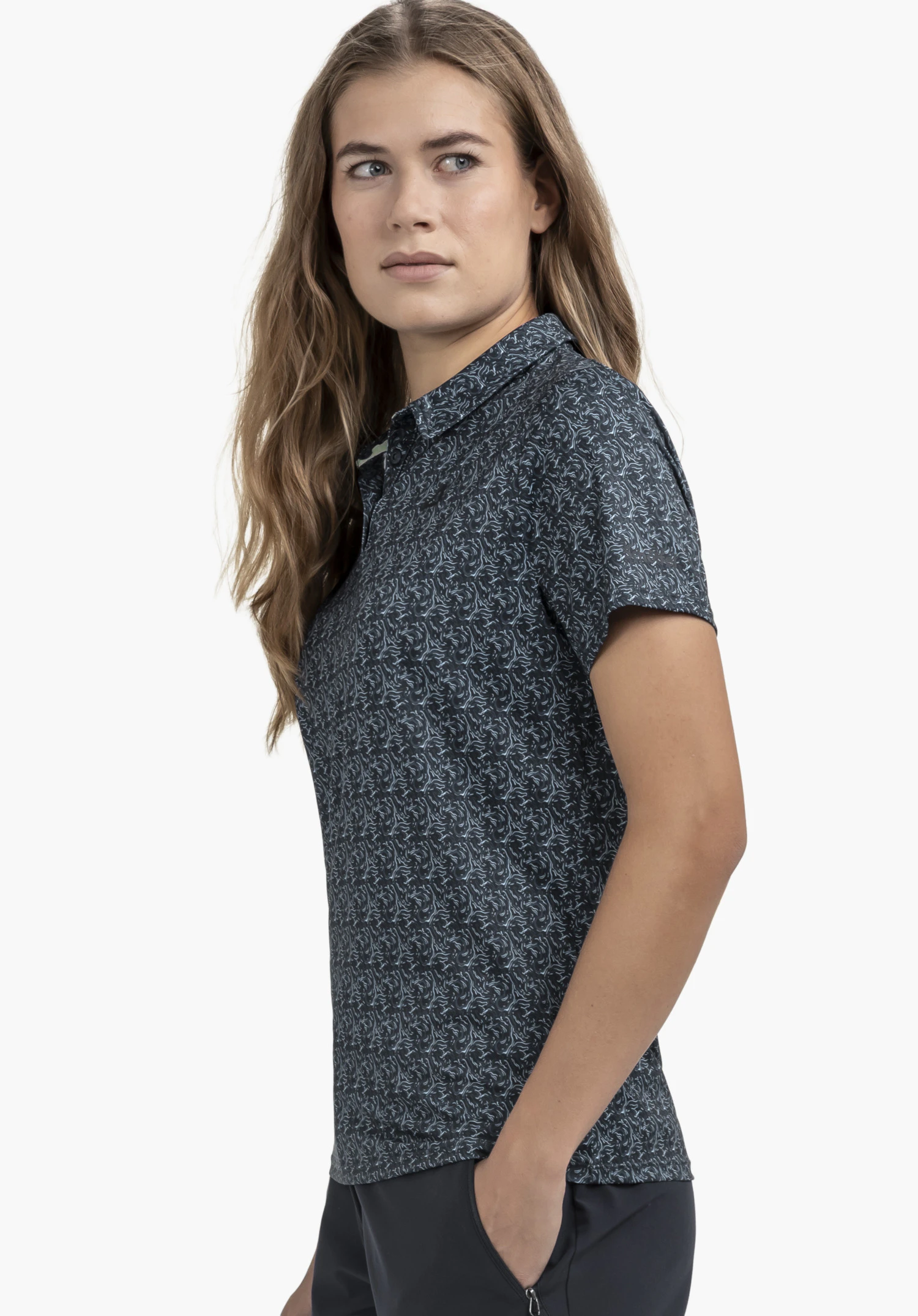 Polo Shirt Style Fraydo WMN in Blau präsentiert von einer Frau, die seitlich steht und in die Kamera blickt. Das Oberteil ist vor einem neutralen Hintergrund zu sehen, das dezente Schöffel-Logo auf dem Ärmel unterstreicht den sportlichen Look.