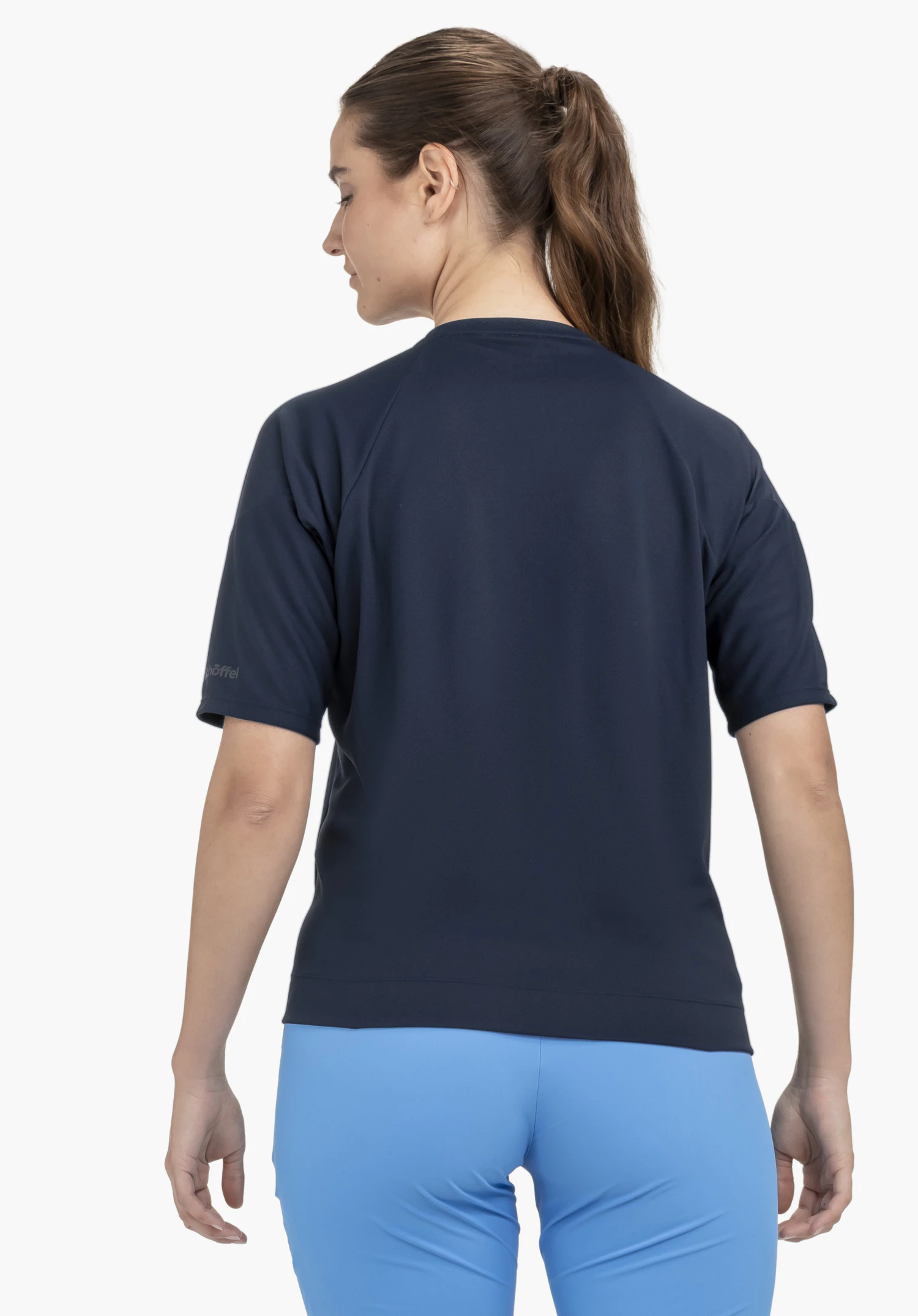 T Shirt Style Enixa WMN getragen von einem Model in der Rückenansicht. Das blaue Shirt zeigt das Schöffel-Logo auf dem linken Ärmel und wird vor einem neutralen Hintergrund präsentiert.