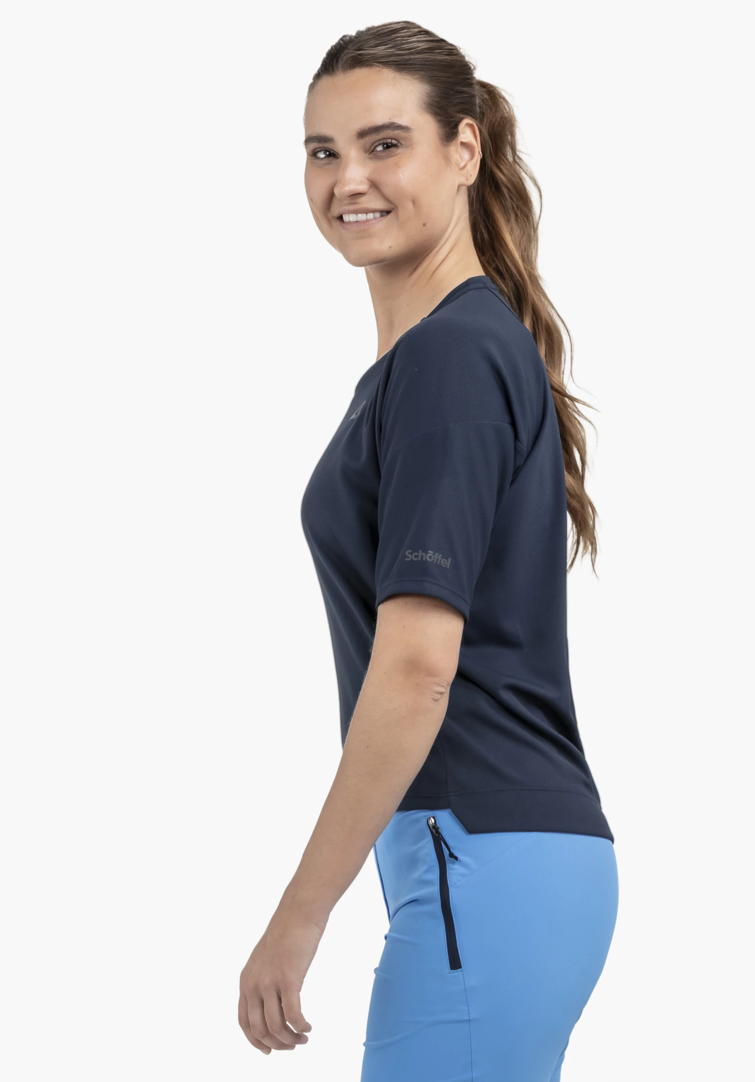 T Shirt Style Enixa WMN: Das Model steht seitlich vor einem neutralen Hintergrund und präsentiert das Shirt in elegantem Blau. Das dezent platzierte Schöffel-Logo am Ärmel unterstreicht den sportiven Look für die kommende Saison.