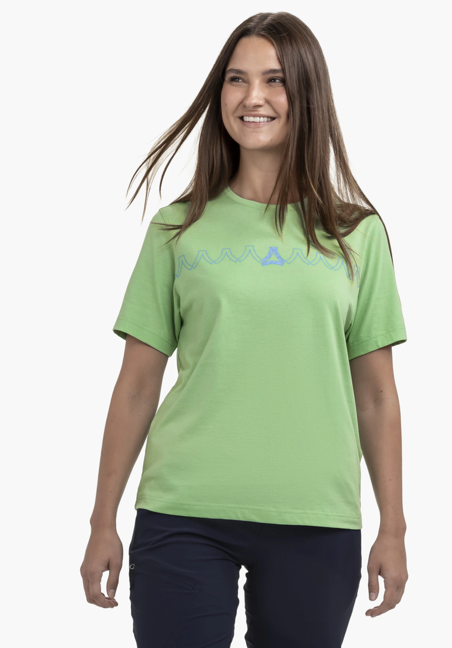T Shirt Style Elmori WMN, getragen von einem Model, das frontal steht, lächelt und leicht zur Seite blickt. Das grüne Shirt zeigt ein grafisches Muster mit dem Schöffel-Logo auf der Brust und wird vor einem neutralen Hintergrund präsentiert.