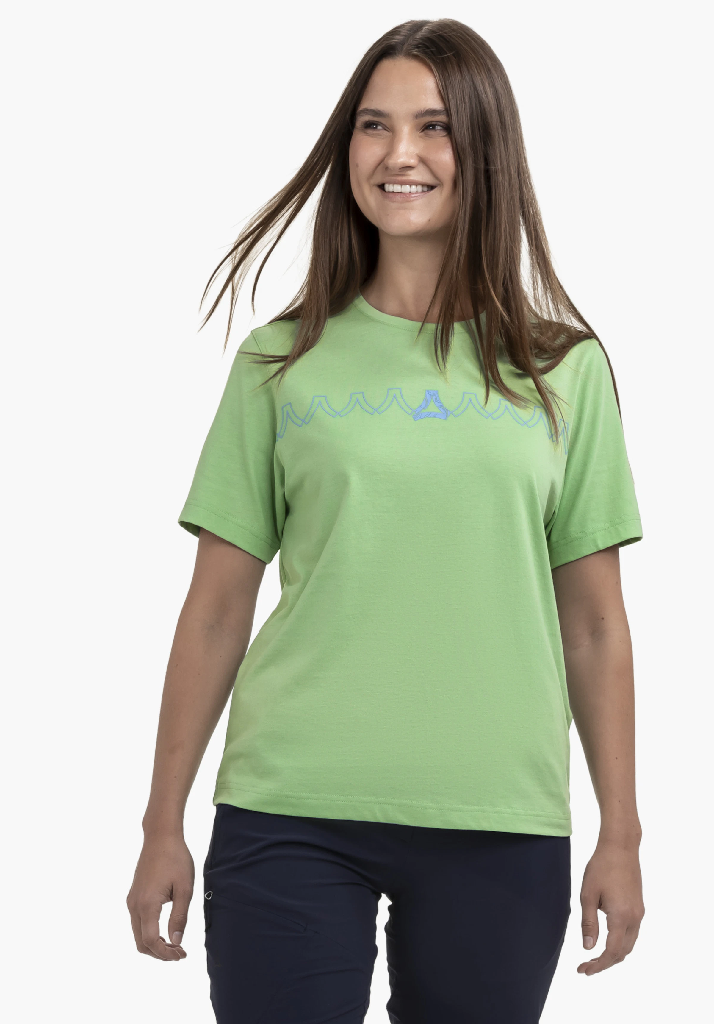 T Shirt Style Elmori WMN, worn by a model standing frontally, smiling and looking slightly to the side. The green shirt features a graphic pattern with the Schöffel-Logo on the chest and is presented against a neutral background.