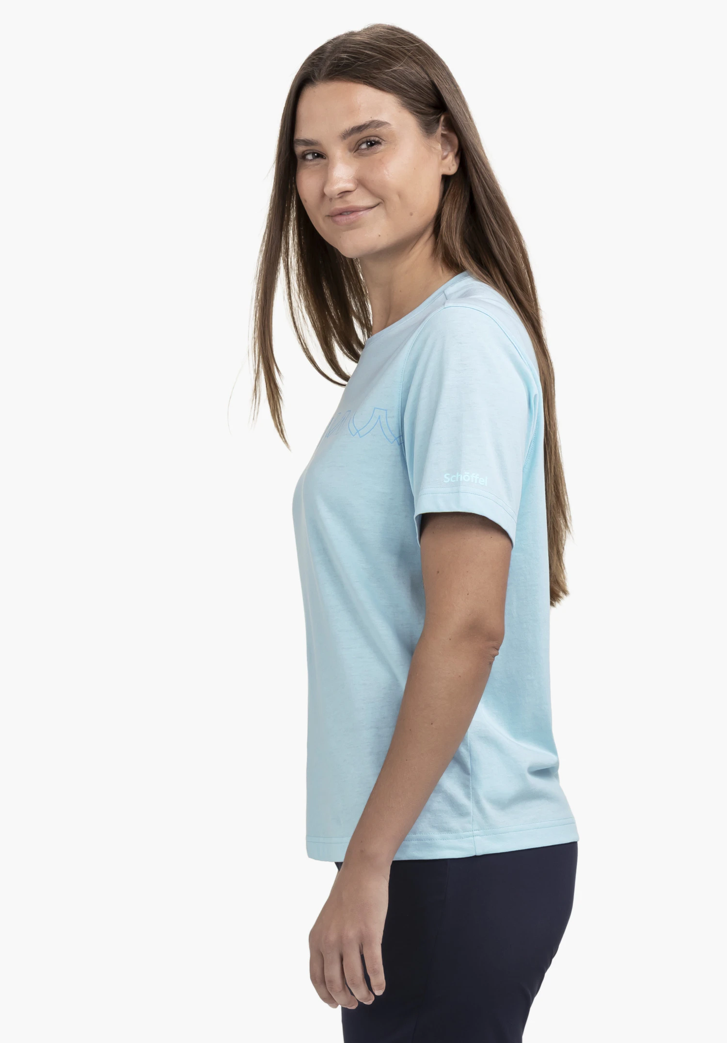 T Shirt Style Elmori WMN bleu porté par une femme souriante, vue de profil. Le mannequin se tient devant un fond neutre et met en valeur le discret logo Schöffel sur la manche.