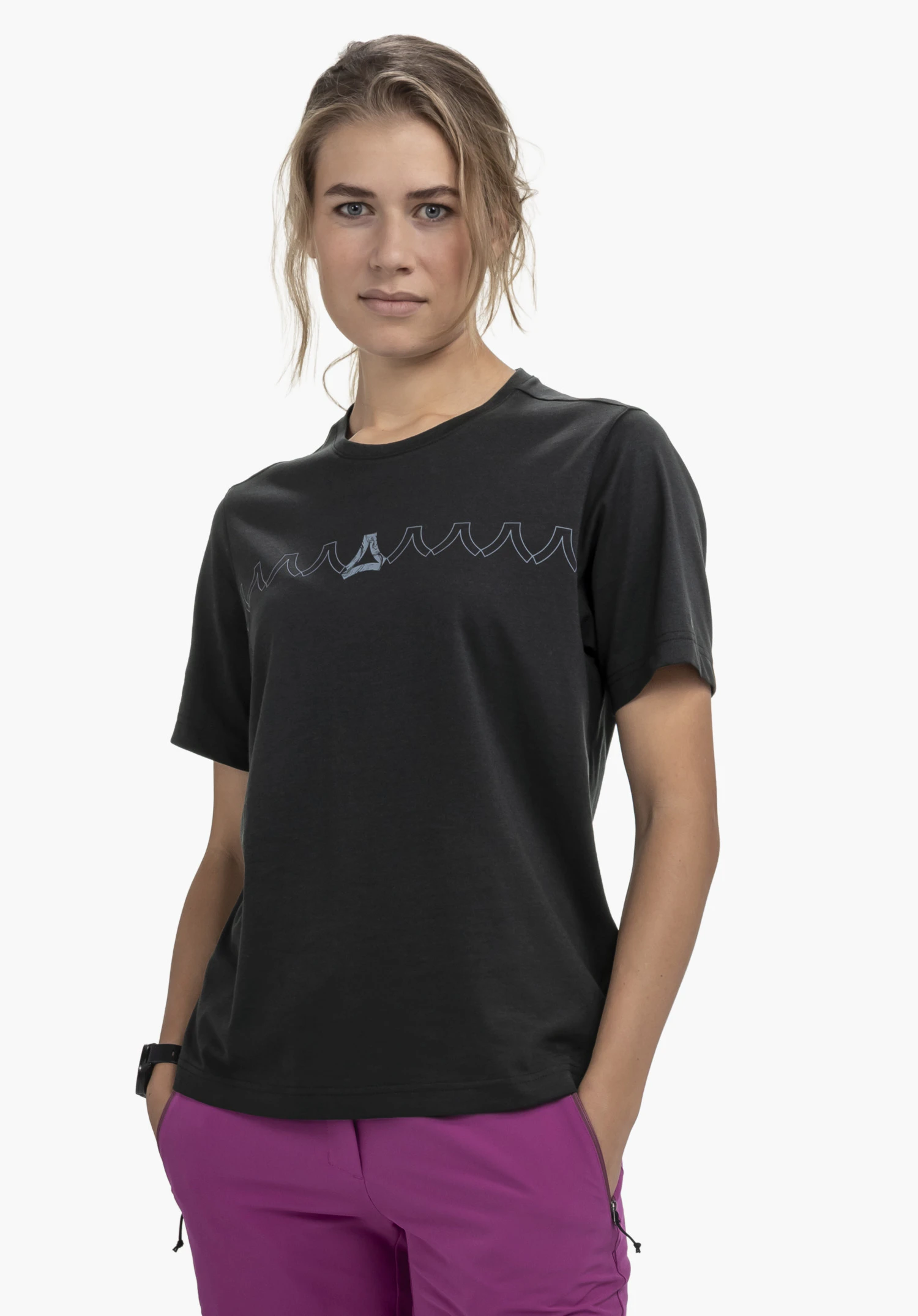 T Shirt Style Elmori WMN in black, worn by a female model standing relaxed with her hands in the pockets of her pink trousers in front of a white background. A graphic print featuring the Schöffel logo is visible on the chest.