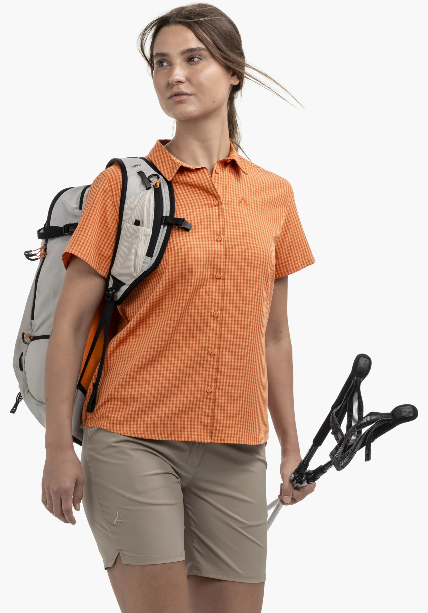 CIRC Blouse Style Dalfoss WMN. Un mannequin se tient légèrement de profil et présente le chemisier à manches courtes à carreaux orange avec un discret logo Schöffel sur la poitrine. Elle porte un sac à dos et tient des bâtons de marche devant un fond neutre.