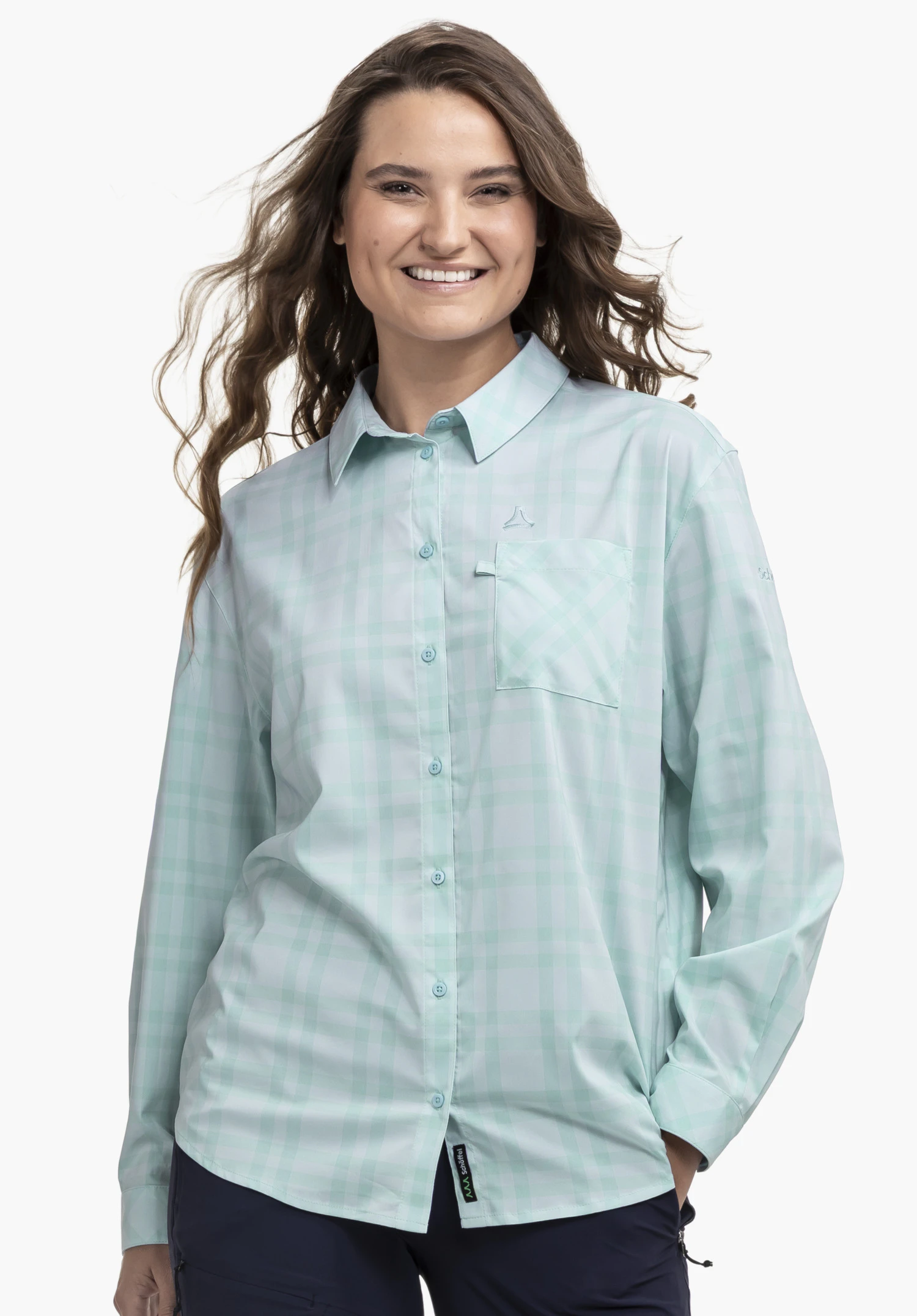 Blouse Style Fraris WMN worn by a model standing frontally and smiling at the camera. The worn view shows the long-sleeved, chequered blouse in blue with a chest pocket and the visible Schöffel logo on the left chest against a neutral background.
