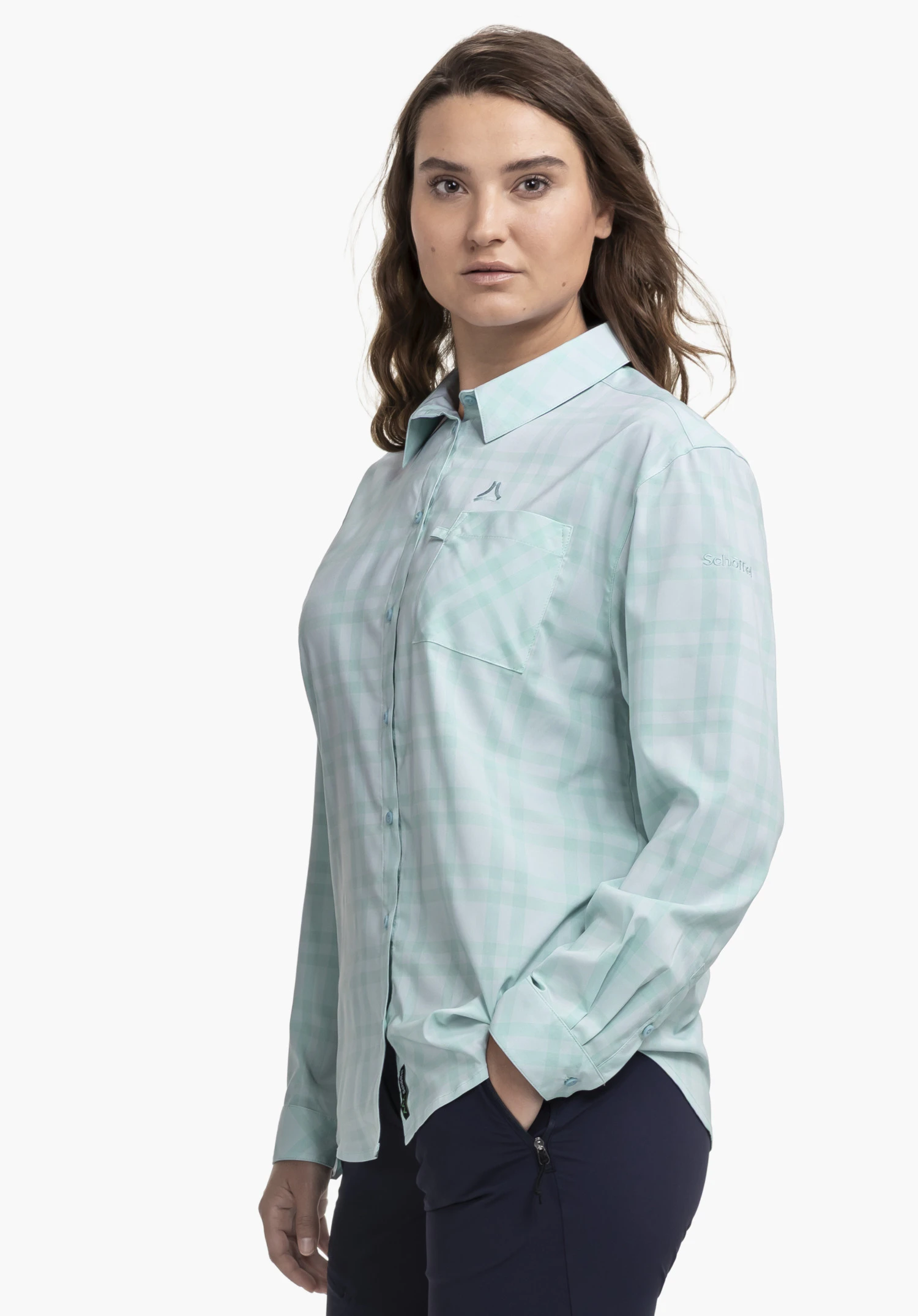 Blouse Style Fraris WMN worn by a model standing slightly to the side. The long-sleeved, chequered blouse in blue features a chest pocket with the Schöffel logo visible above it. Another Schöffel logo is located on the left sleeve. The image is set against a neutral background.