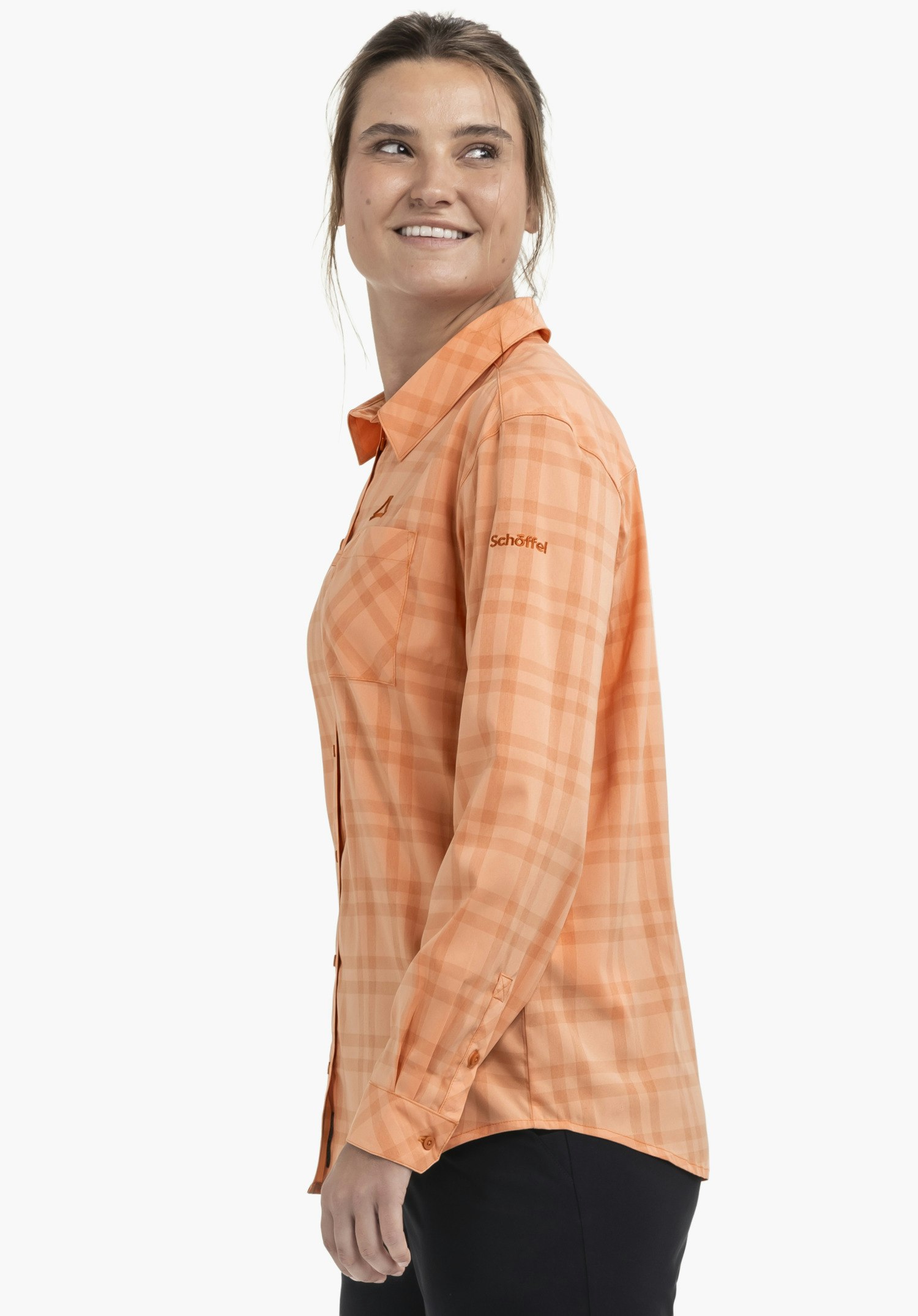 Blouse Style Fraris WMN portée par un mannequin. Le chemisier à carreaux orange présente le Schöffel-Logo sur la manche gauche. Le mannequin se tient de profil et sourit devant un fond neutre.
