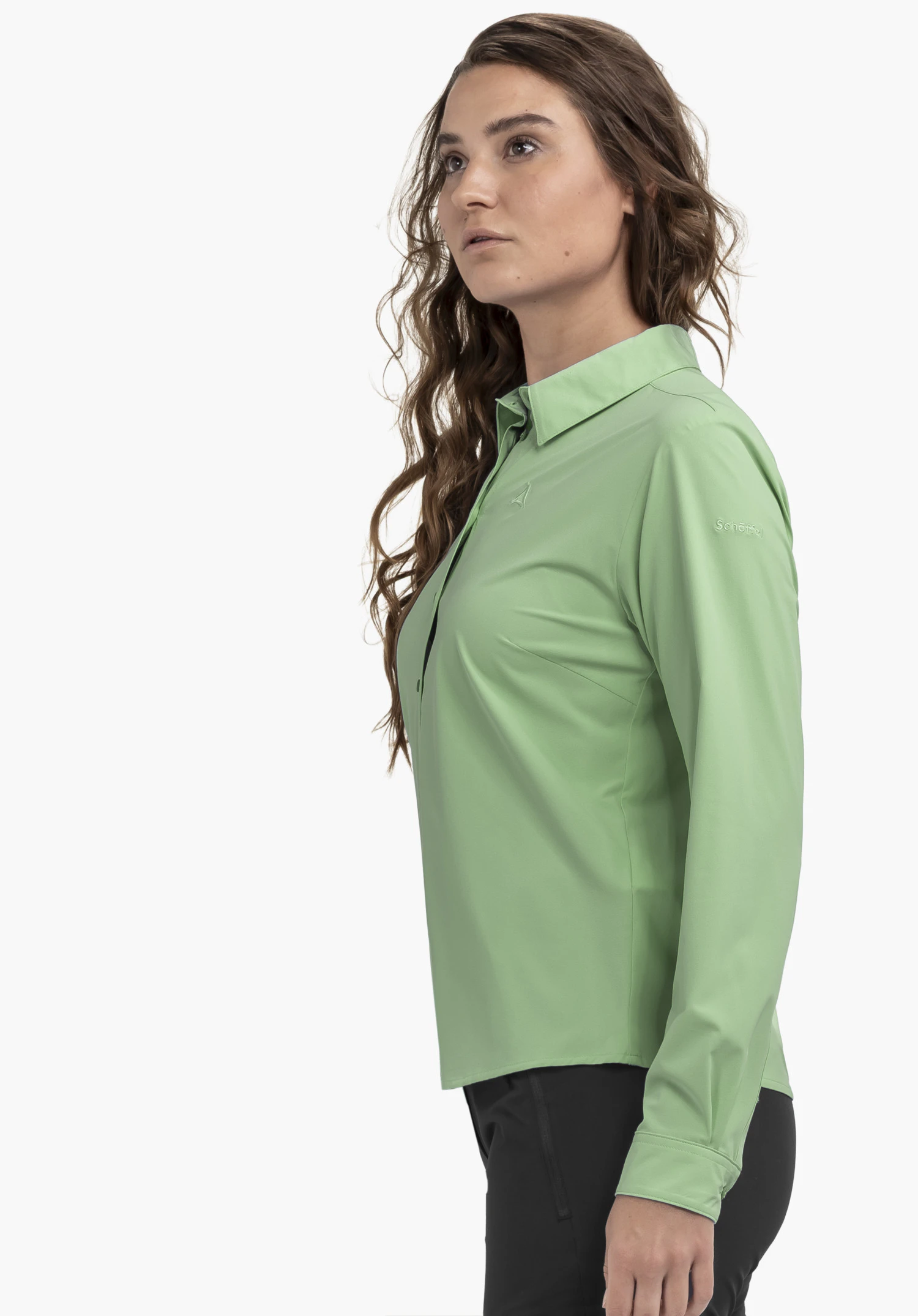 Blouse Style Dunajec WMN zeigt eine Frau, die seitlich vor einem neutralen Hintergrund steht. Die Bluse in Grün betont den Schnitt, während das dezente Schöffel-Logo auf dem Oberarm für Qualität steht.