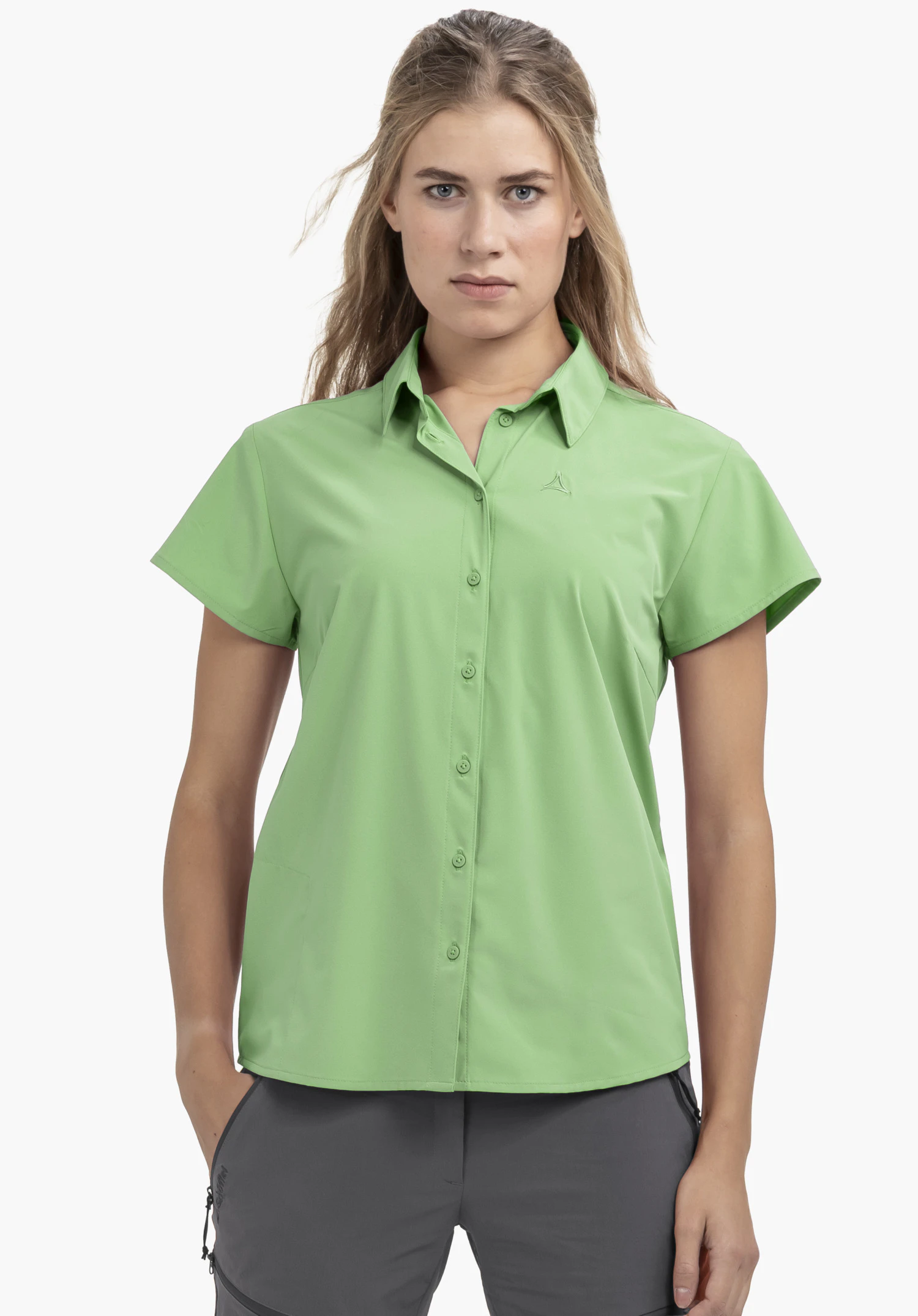Blouse Style Dooser WMN portée par un mannequin se tenant de face, la main droite dans la poche de son pantalon. Le chemisier vert à manches courtes présente un discret logo Schöffel sur la poitrine gauche et est présenté sur un fond neutre.