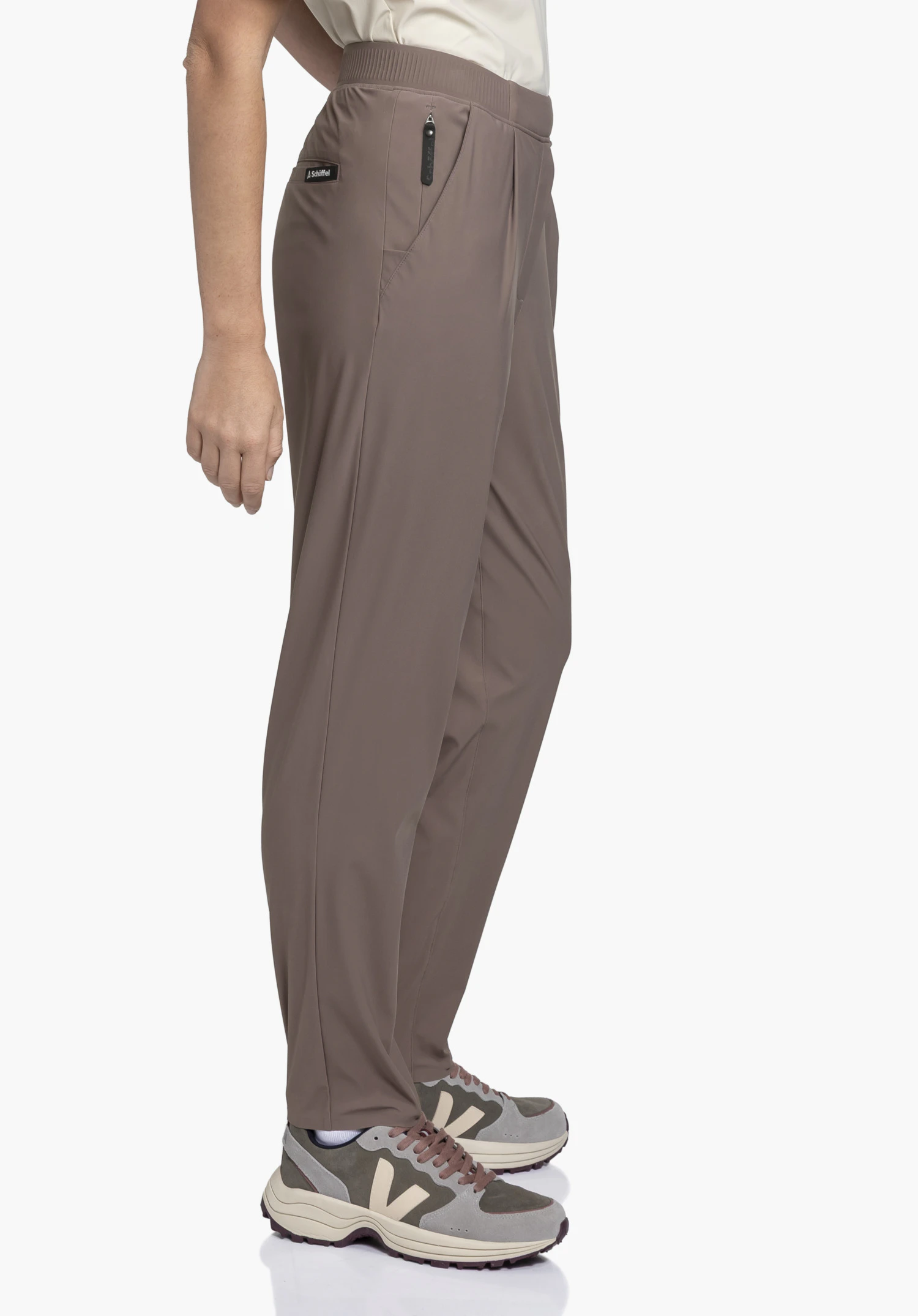 Jersey Pants Style Calmay WMN présentée dans un coloris marron élégant. Le mannequin pose de profil devant un arrière-plan neutre, mettant en valeur la coupe du pantalon, avec le logo Schöffel discrètement visible sur la ceinture.