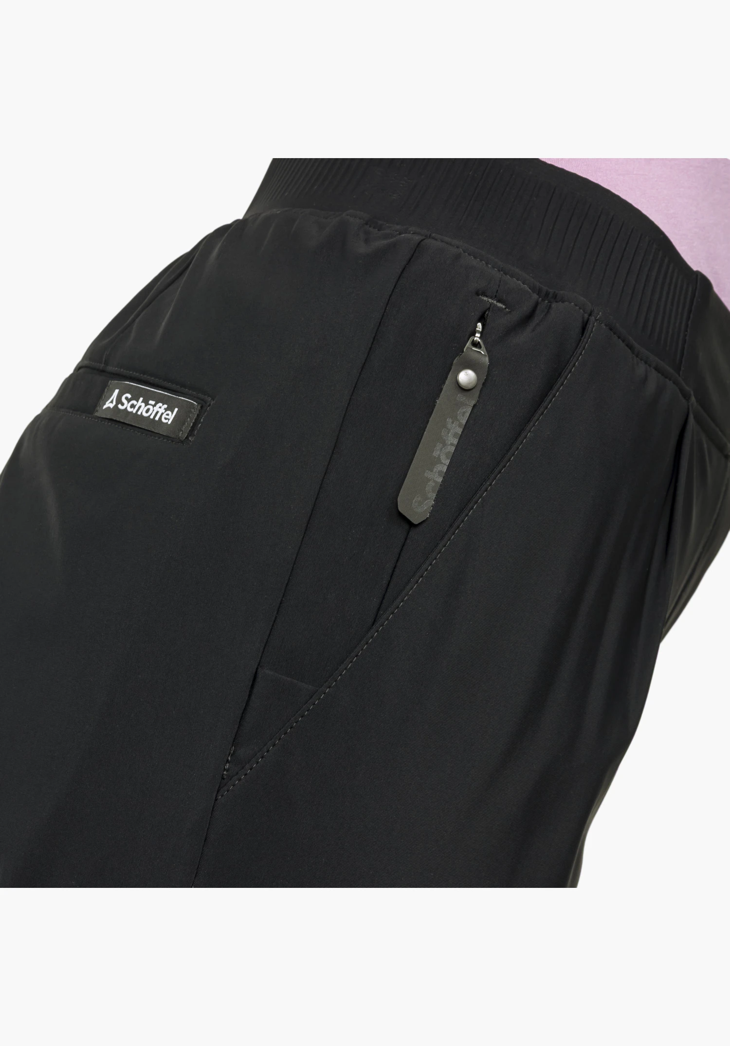 Jersey Pants Style Calmay WMN montrant les détails du tissu noir, une poche latérale zippée avec une tirette distinctive et un petit écusson du logo Schöffel, gros plan sur fond blanc/neutre.