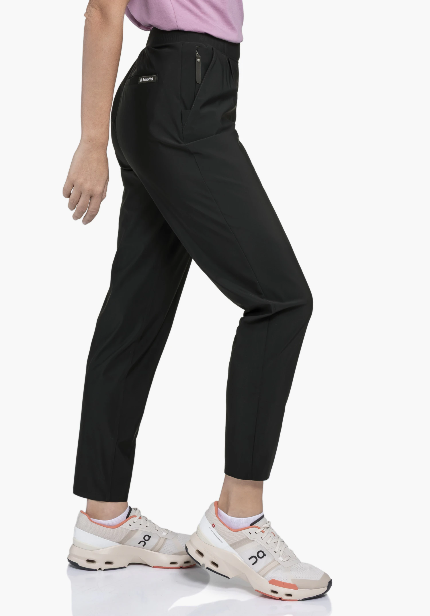 Jersey Pants Style Calmay WMN sur un mannequin en vue latérale. Le mannequin se tient de profil et porte le pantalon noir, sur lequel le logo Schöffel est visible sous la ceinture, devant un fond neutre.