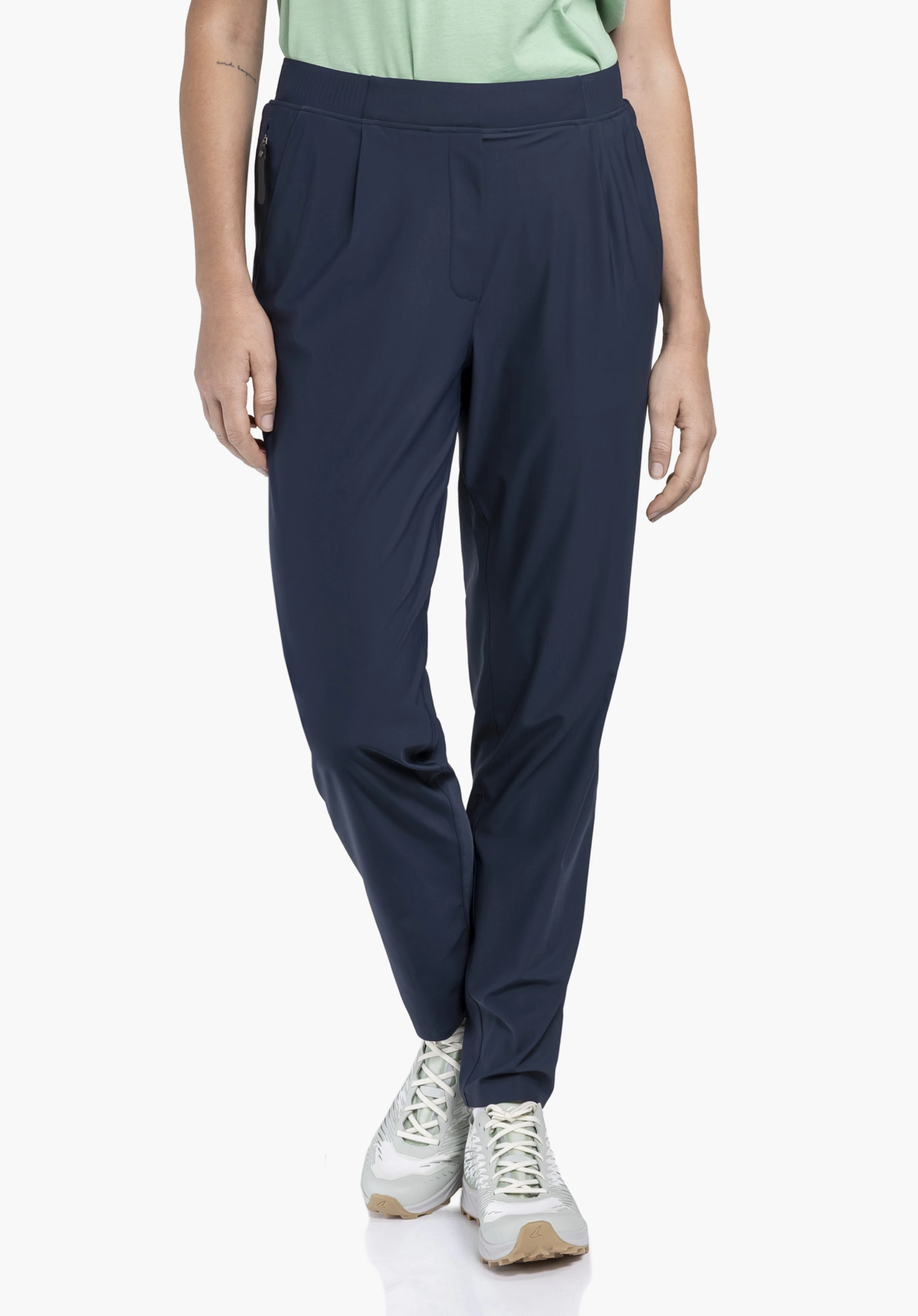Jersey Pants Style Calmay WMN portée par un mannequin de face. Elle porte le pantalon bleu avec d'élégants plis à l'avant et des chaussures de sport devant un fond neutre.