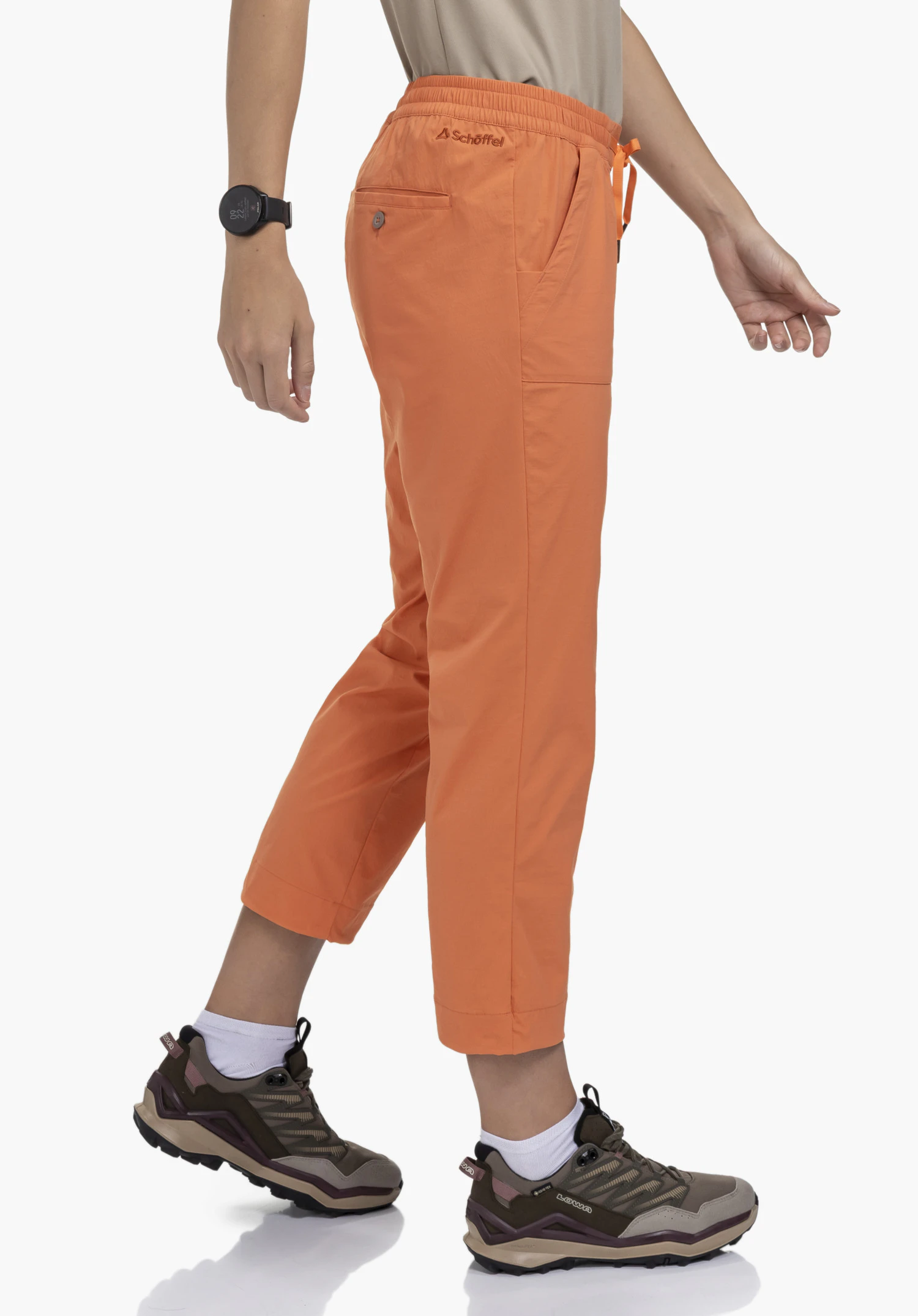 Pants Rangun L en orange, portée par un mannequin debout de profil dans une posture de marche. Le logo Schöffel est visible à l'arrière sous la ceinture, présenté sur un fond neutre.