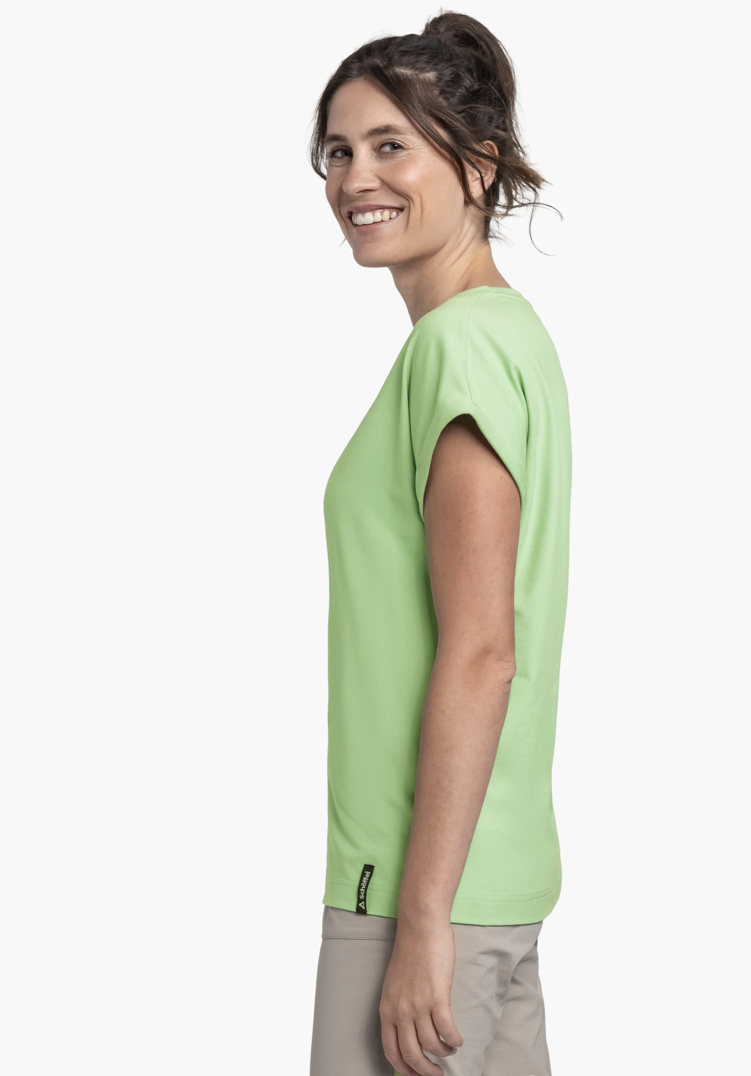 T Shirt Style Serenio WMN in green, worn by a smiling model standing sideways. A small Schöffel-Logo is placed on the bottom hem, shot against a neutral background.