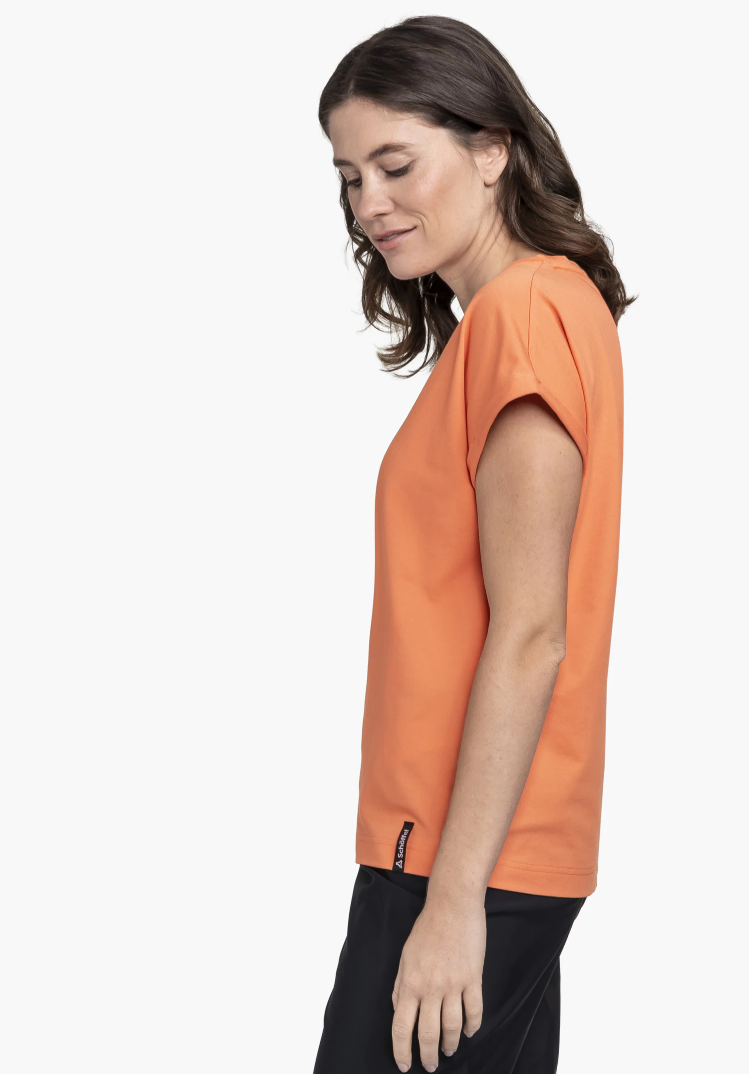 T Shirt Style Serenio WMN in orange. A model stands sideways wearing the shirt with a small Schöffel-Logo on the lower side hem against a neutral background.