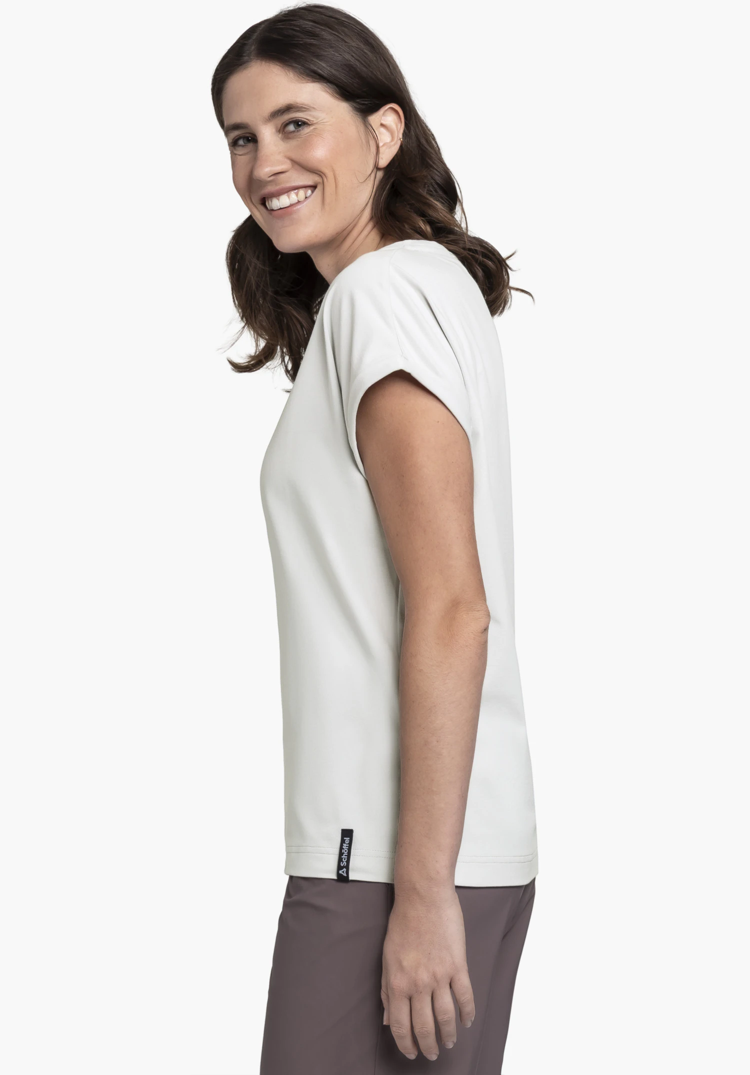 T Shirt Style Serenio WMN en gris. Un mannequin femme porte le t-shirt, se tient de profil et regarde l'objectif en souriant. Un petit logo Schöffel est visible sur l'ourlet inférieur, photographié devant un fond neutre.