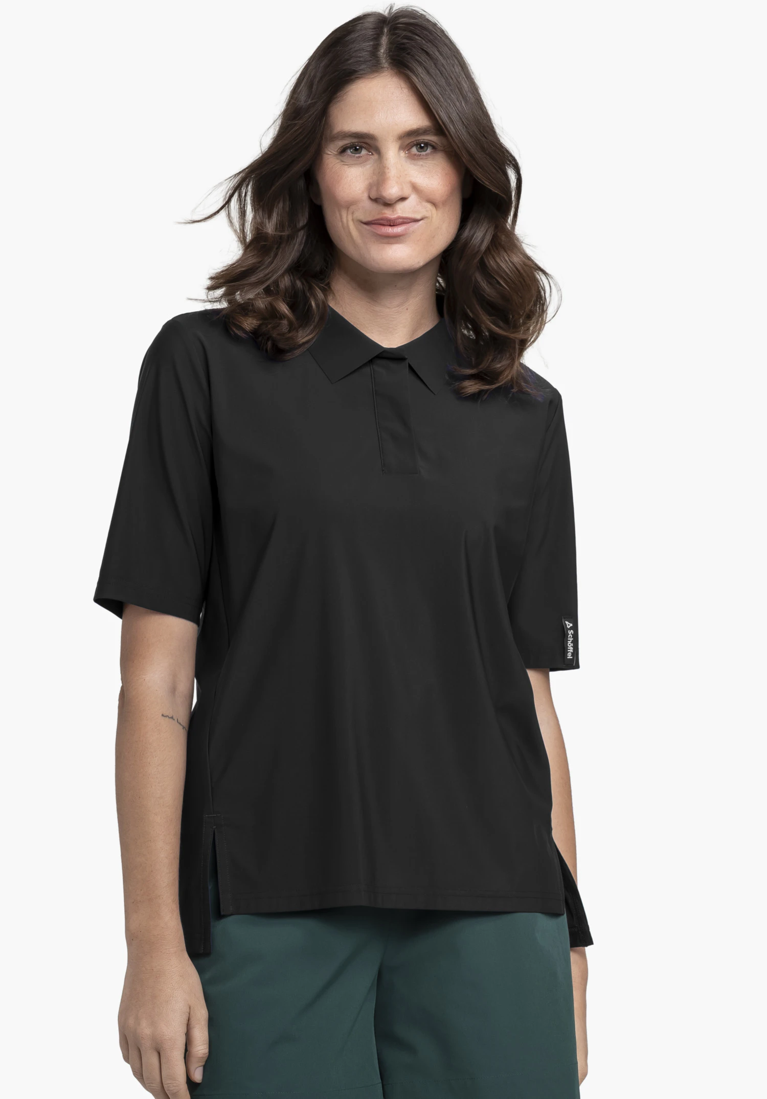 Jersey Polo Style Calmay WMN est porté par un mannequin se tenant de face. Le polo noir présente des manches courtes, un col classique et un logo Schöffel sur l'ourlet de la manche gauche. L'image est présentée sur un fond neutre.