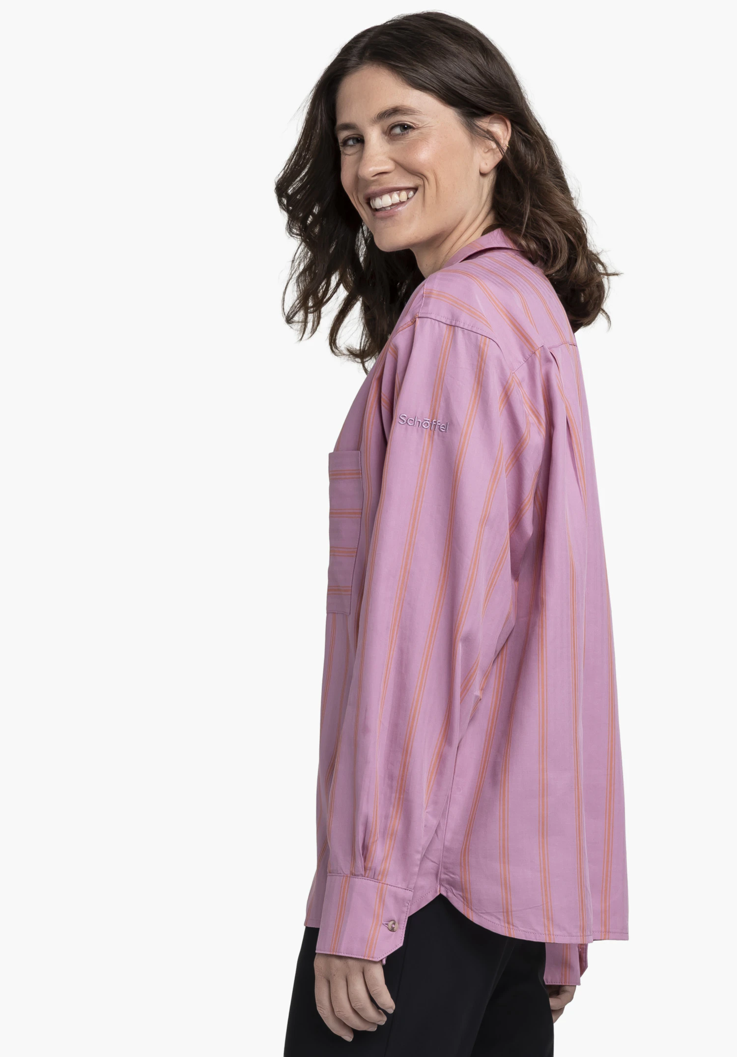 Blouse Style Chanduy WMN sur un mannequin se tenant de profil et souriant par-dessus son épaule à l'appareil photo. Le chemisier à manches longues rose présente un fin motif à rayures et affiche le logo Schöffel sur le haut du bras, présenté sur un fond neutre.