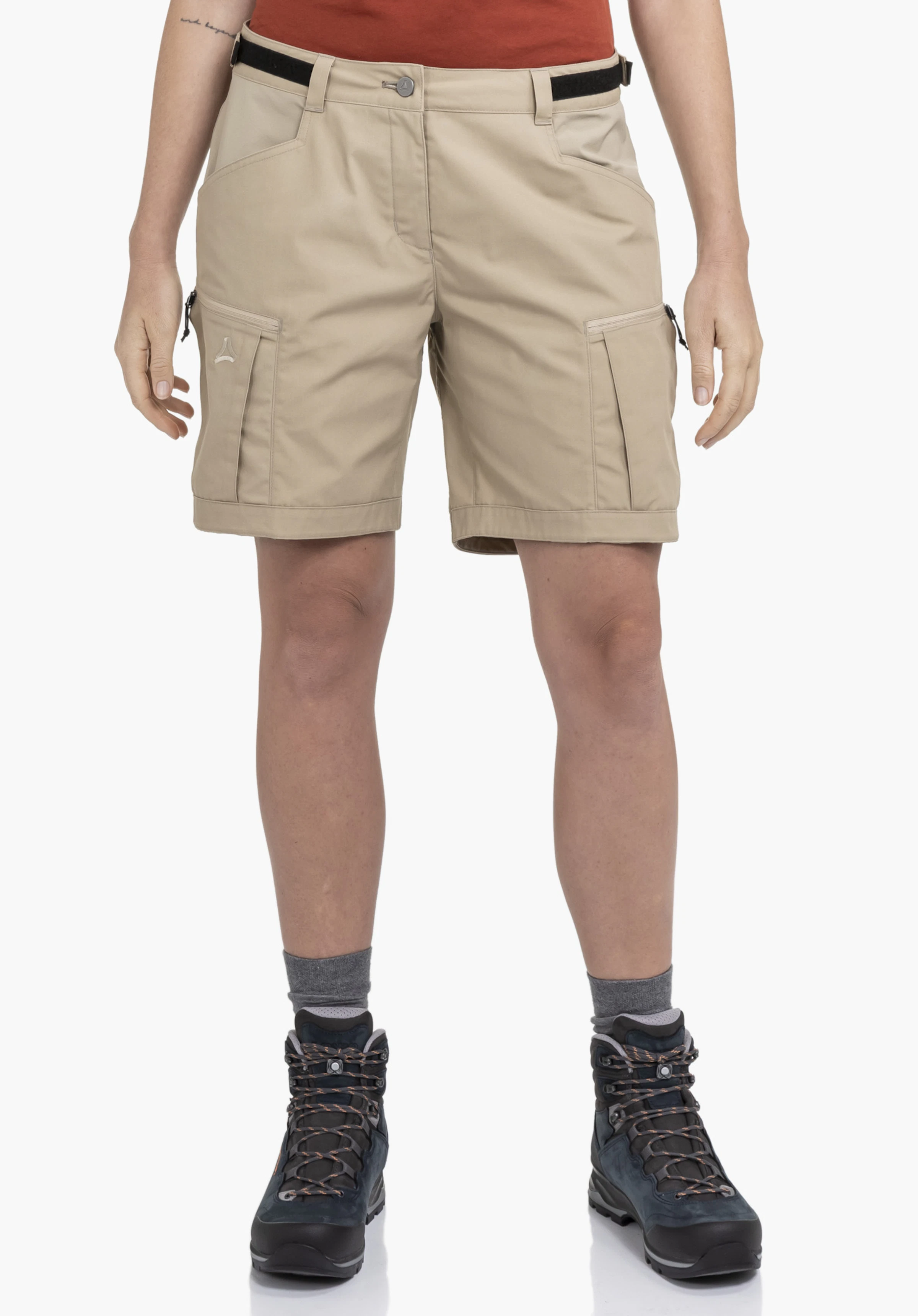 Shorts Style Maghera WMN en beige. Un mannequin se tient de face et porte le short, qui présente des poches cargo latérales avec un logo Schöffel visible sur le côté droit. La tenue est montrée sur un fond neutre, associée à des chaussures de randonnée.