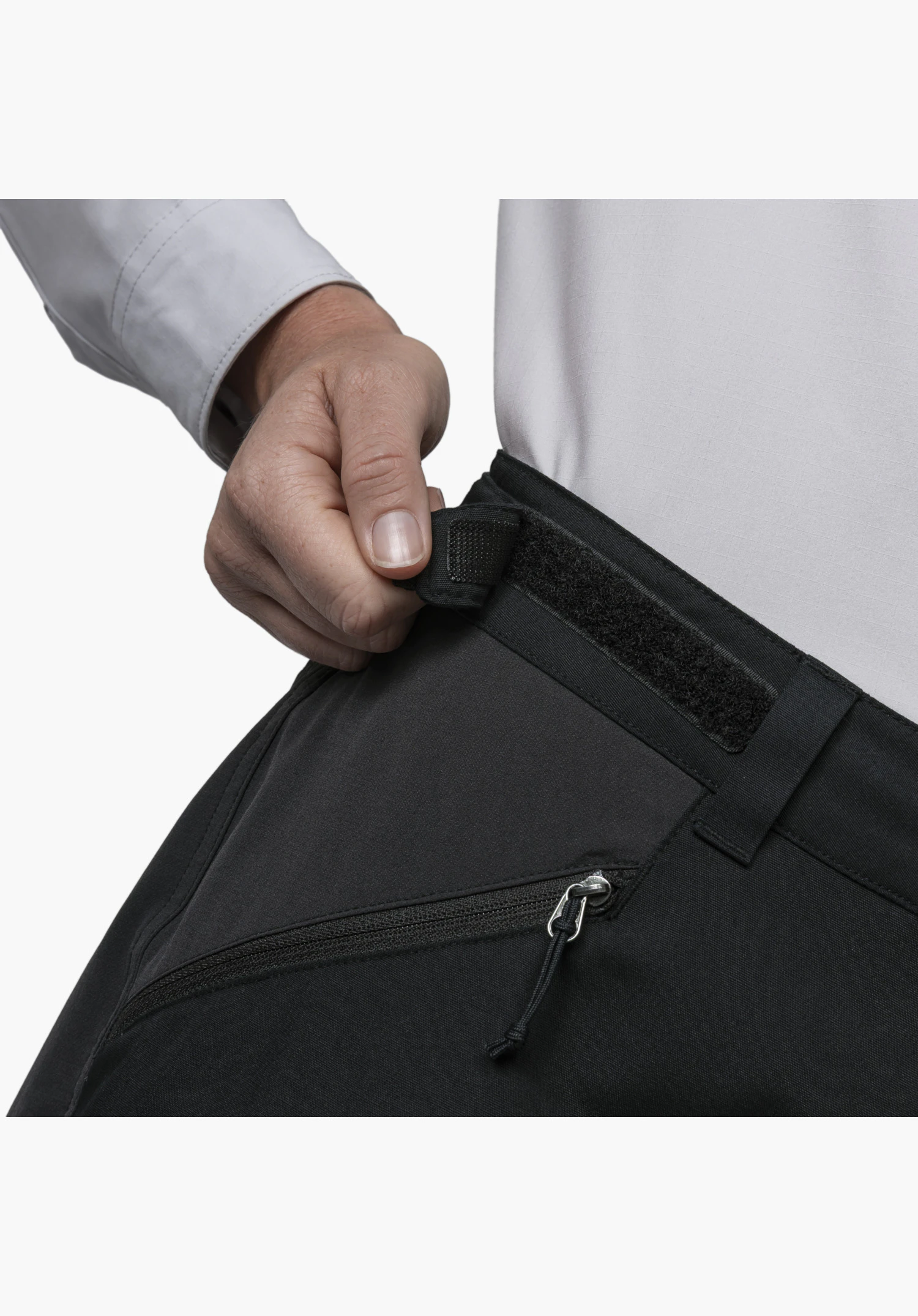 Pants Style Maghera WMN montre une main ajustant la fermeture auto-agrippante noire de la ceinture, située au-dessus d'une poche zippée, en gros plan sur fond blanc/neutre.