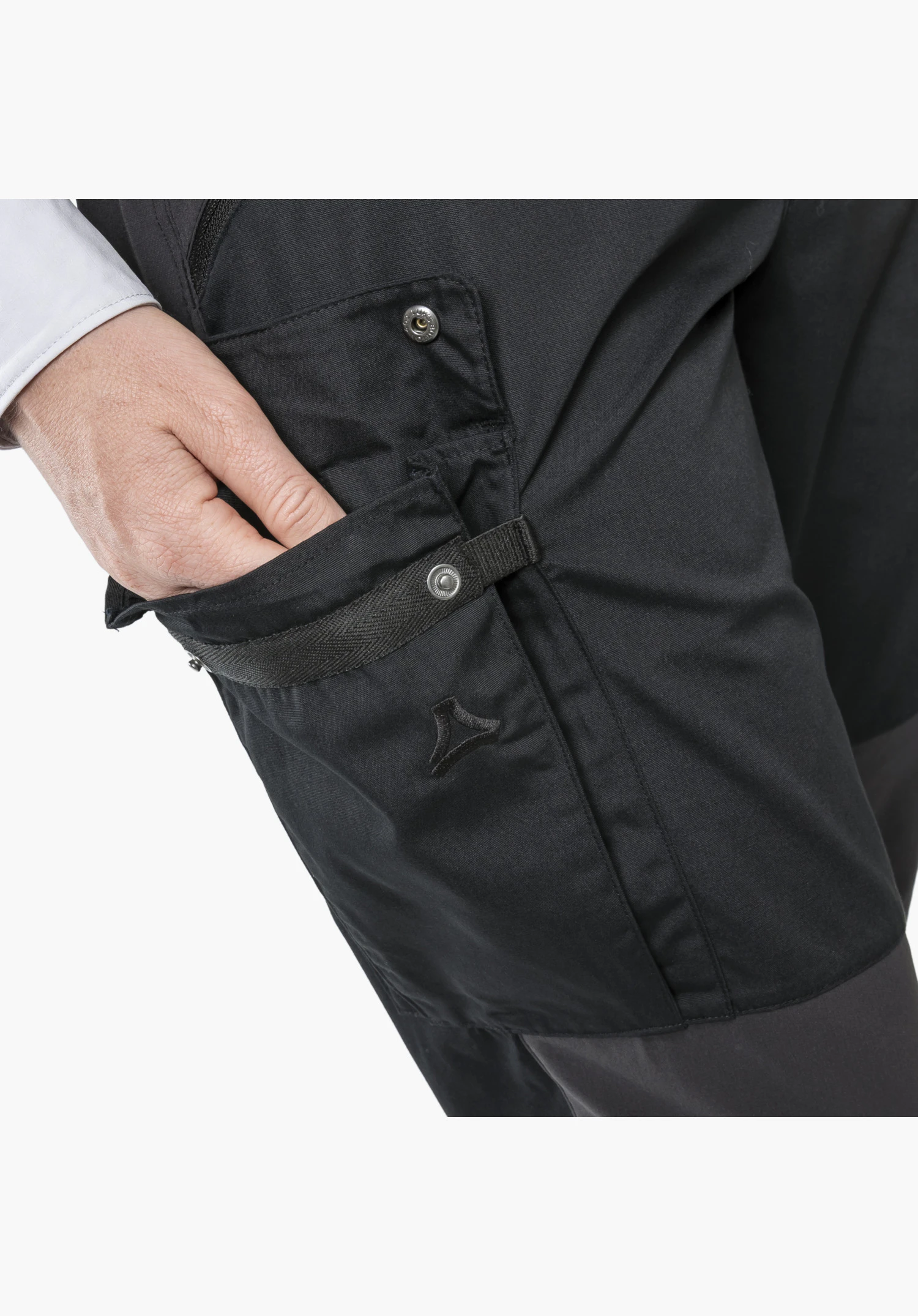 Pants Style Maghera WMN. Une main se glisse dans la poche latérale noire dotée de détails de sangle en tissu, d'un bouton-pression en métal et du logo Schöffel subtilement brodé. Gros plan sur fond blanc.