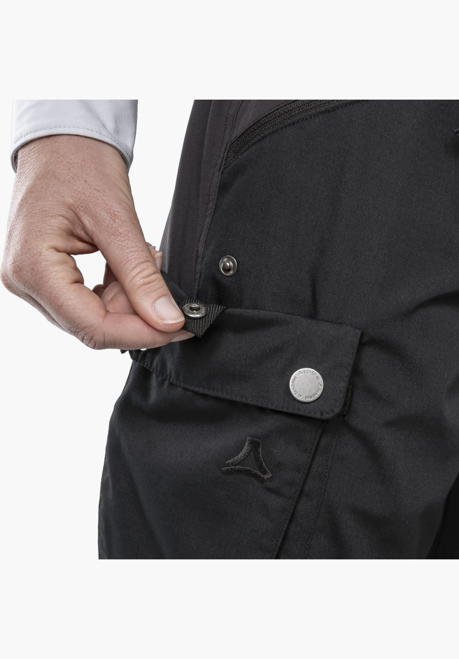 Pants Style Maghera WMN avec un accent sur les détails de la poche. Une main tient une patte de réglage avec bouton-pression sur le tissu noir. Un bouton-pression métallique avec inscription et le logo Schöffel brodé sont clairement visibles, en gros plan sur un fond blanc/neutre.