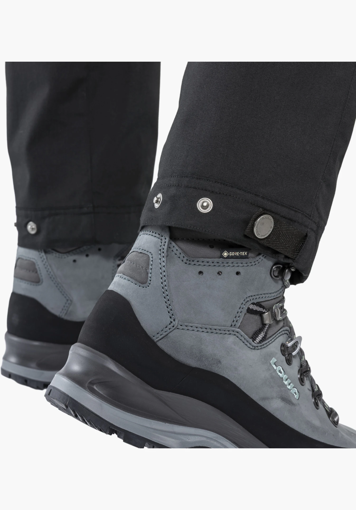 Pants Style Maghera WMN en noir, vue détaillée de l'ourlet de jambe réglable avec des boutons-pression métalliques et le logo Schöffel gravé porté sur une chaussure de randonnée, gros plan sur fond blanc/neutre.