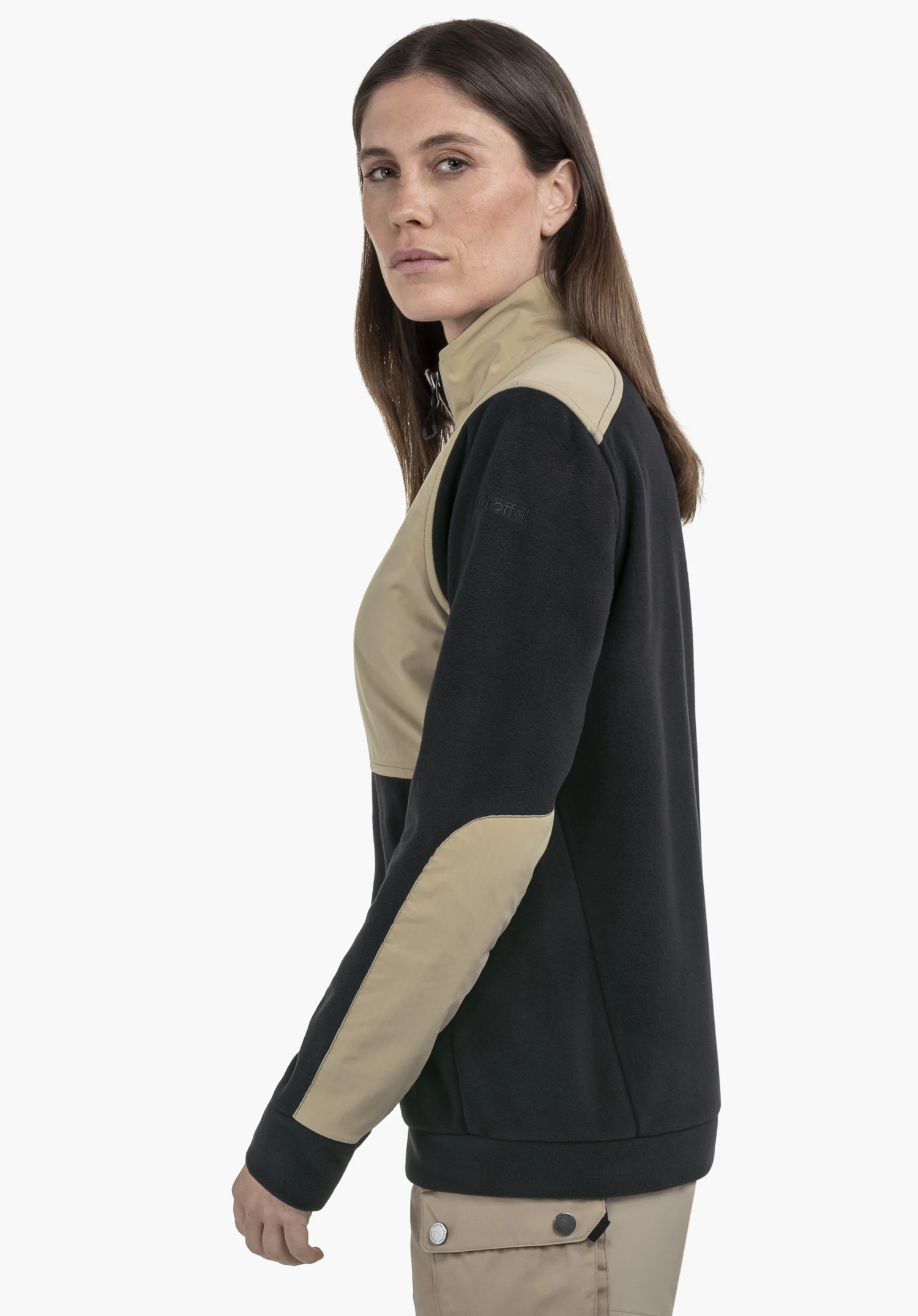 Fleece Jk Style Maghera WMN : Une femme de profil devant un fond neutre. La veste en polaire se distingue par son mélange de couleurs noir et beige. Le logo Schöffel est visible sur le bras, apportant une finition élégante.